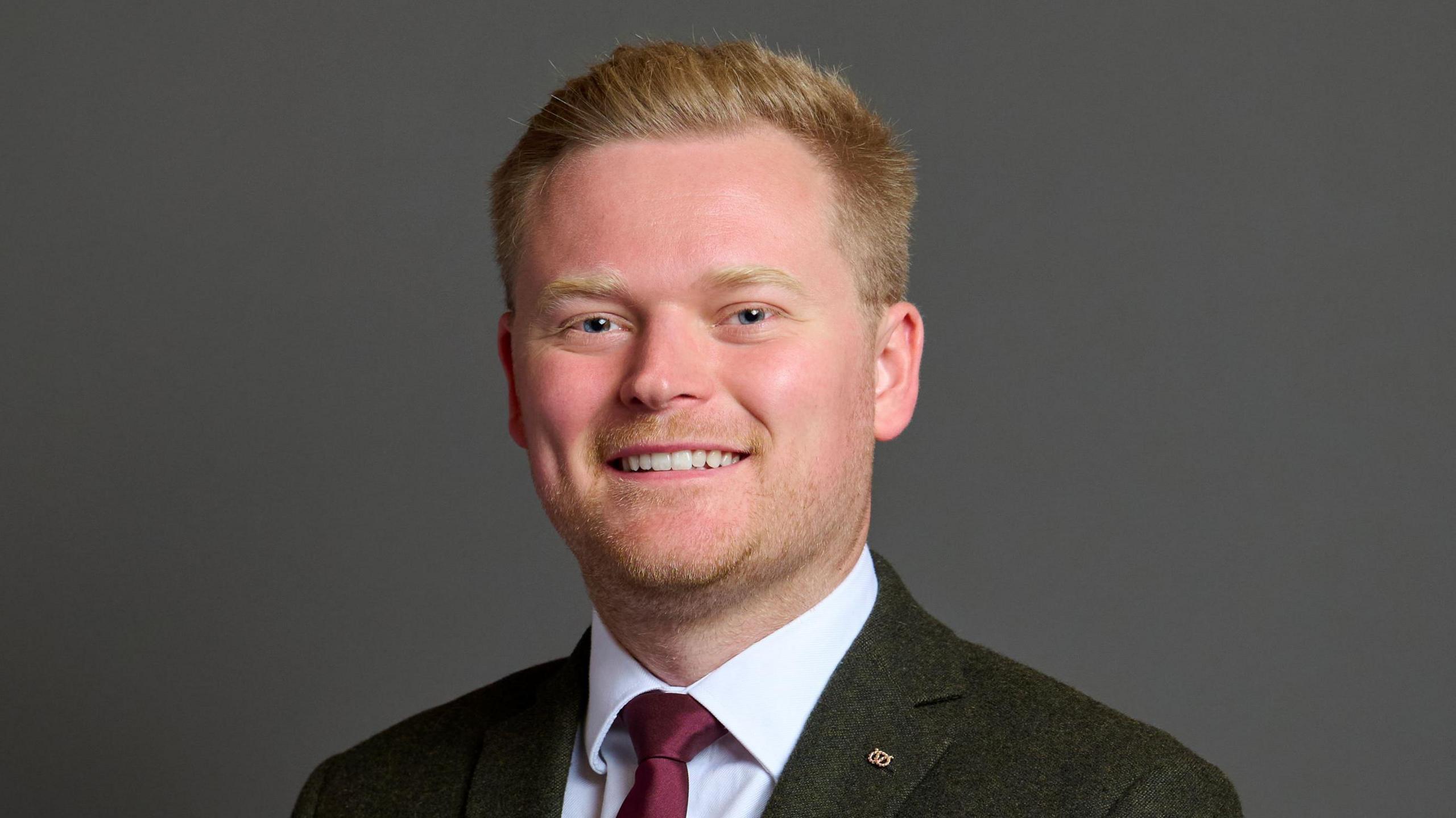 Josh Newbury, a man wearing a dark grey suit and whote shirt with a dark red tie. He is stood against a dark grey background, and is smiling while posing for the camera.