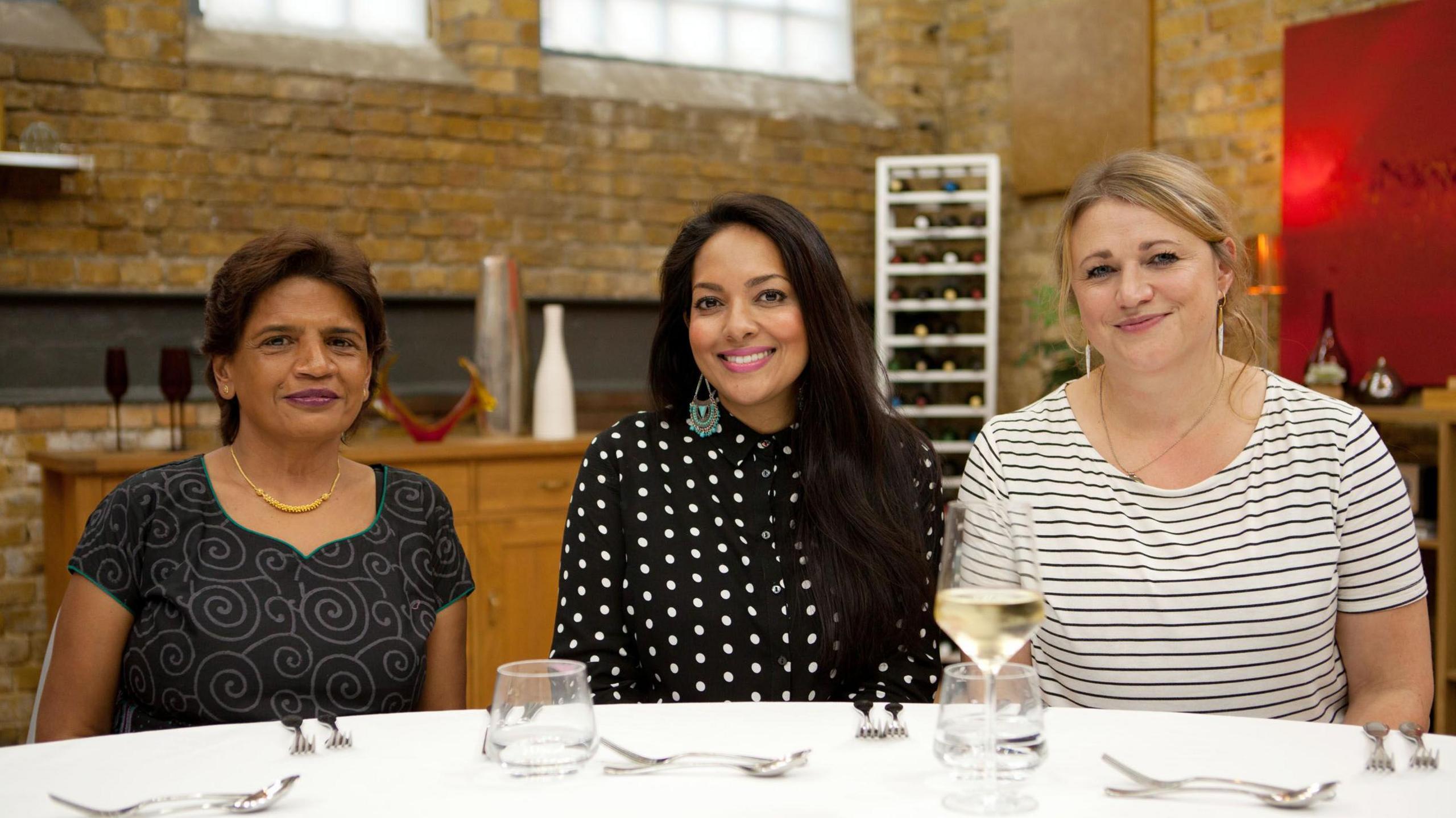 MasterChef guest critics Daksha Mistry, Shelina Permalloo, and Juanita Hennessey. They are smiling and sat at a restaurant table in the studio kitchen.