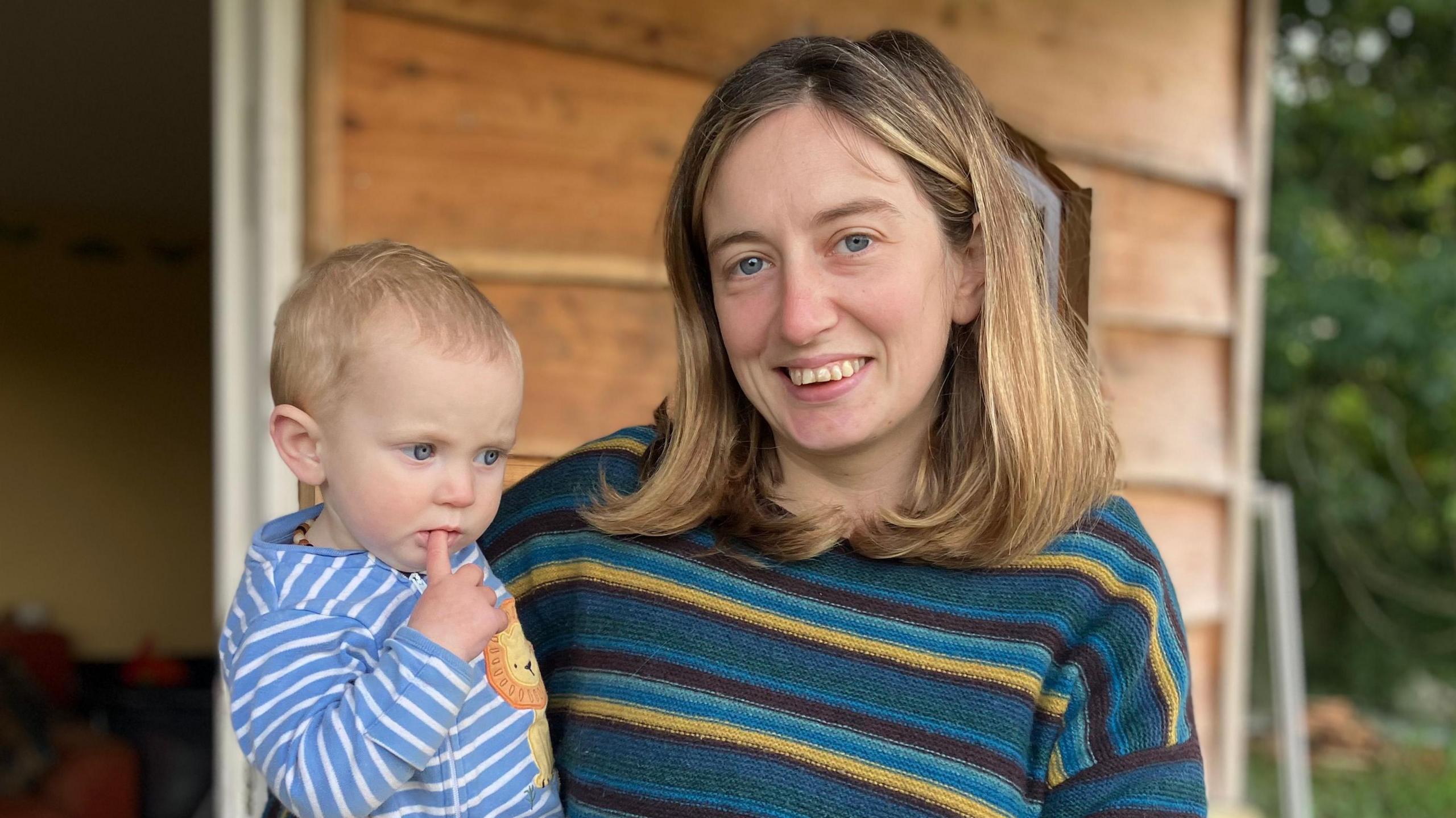 Mimi, who is wearing a teal and yellow striped jumper, is holding her child Nala. Nala is wearing a blue and white striped baby grow which has a cartoon of a lion on.