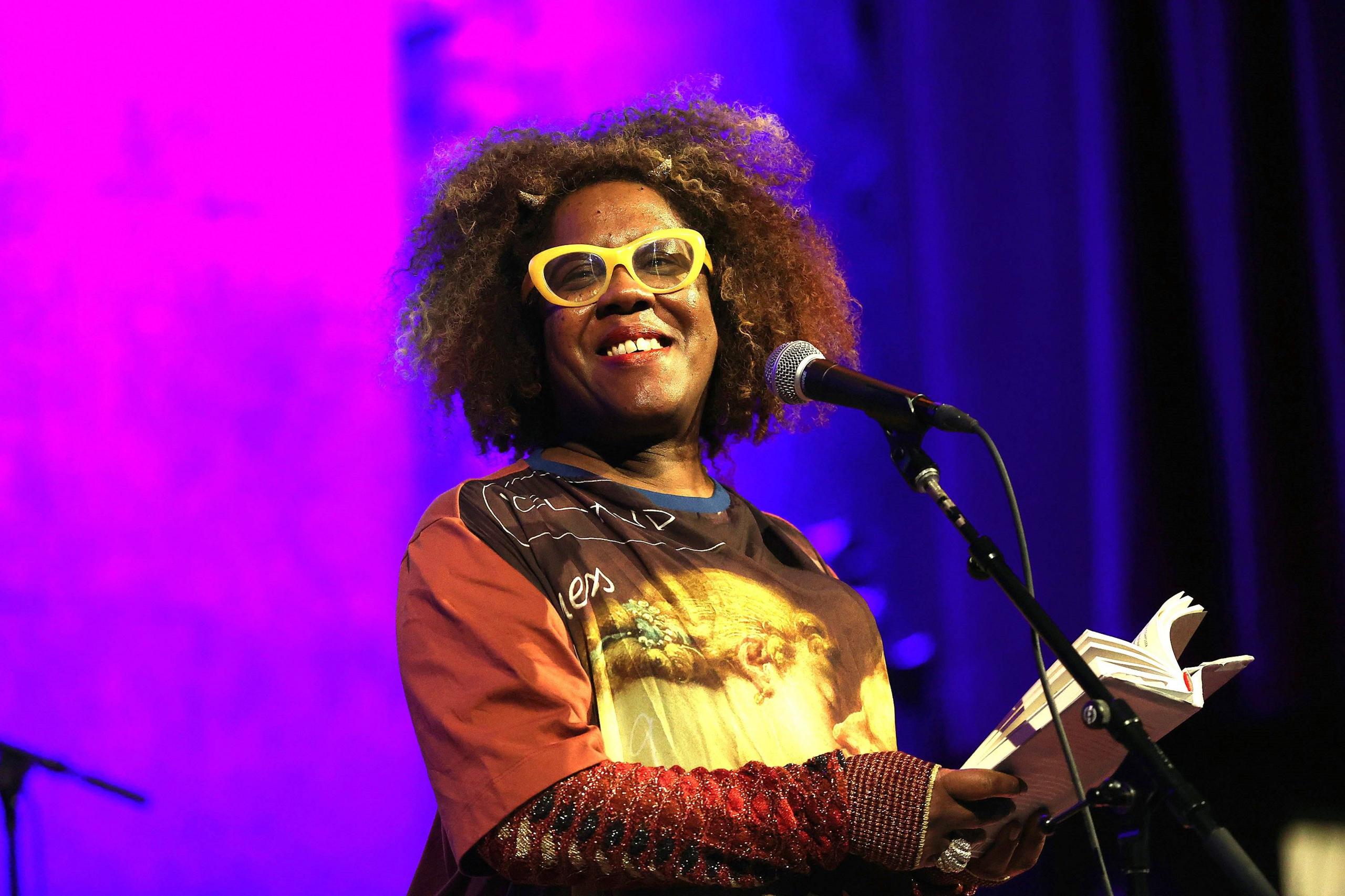 Errollyn Wallen performing in front of a microphone with a book in her hands. She is wearing yellow glasses and a yellow and brown T-shirt over a long-sleeved reddish top. The background is blue and pink.