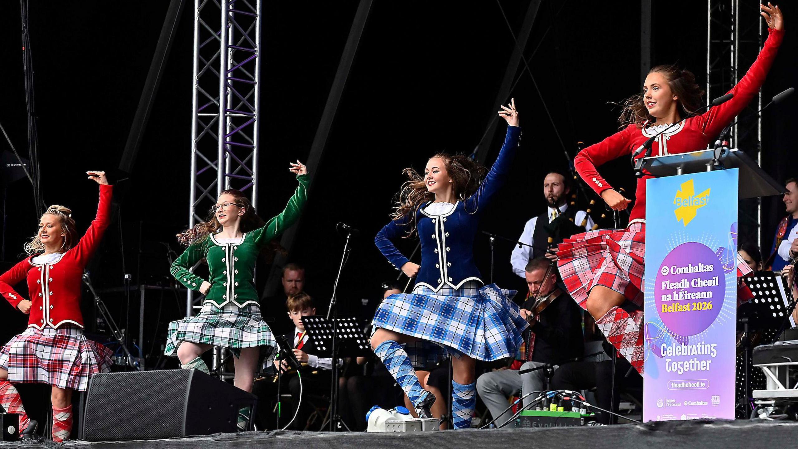Four women in Highland dancing costumes dance on stage in Belfast.   Musicians play instruments including the bagpipes behind them.