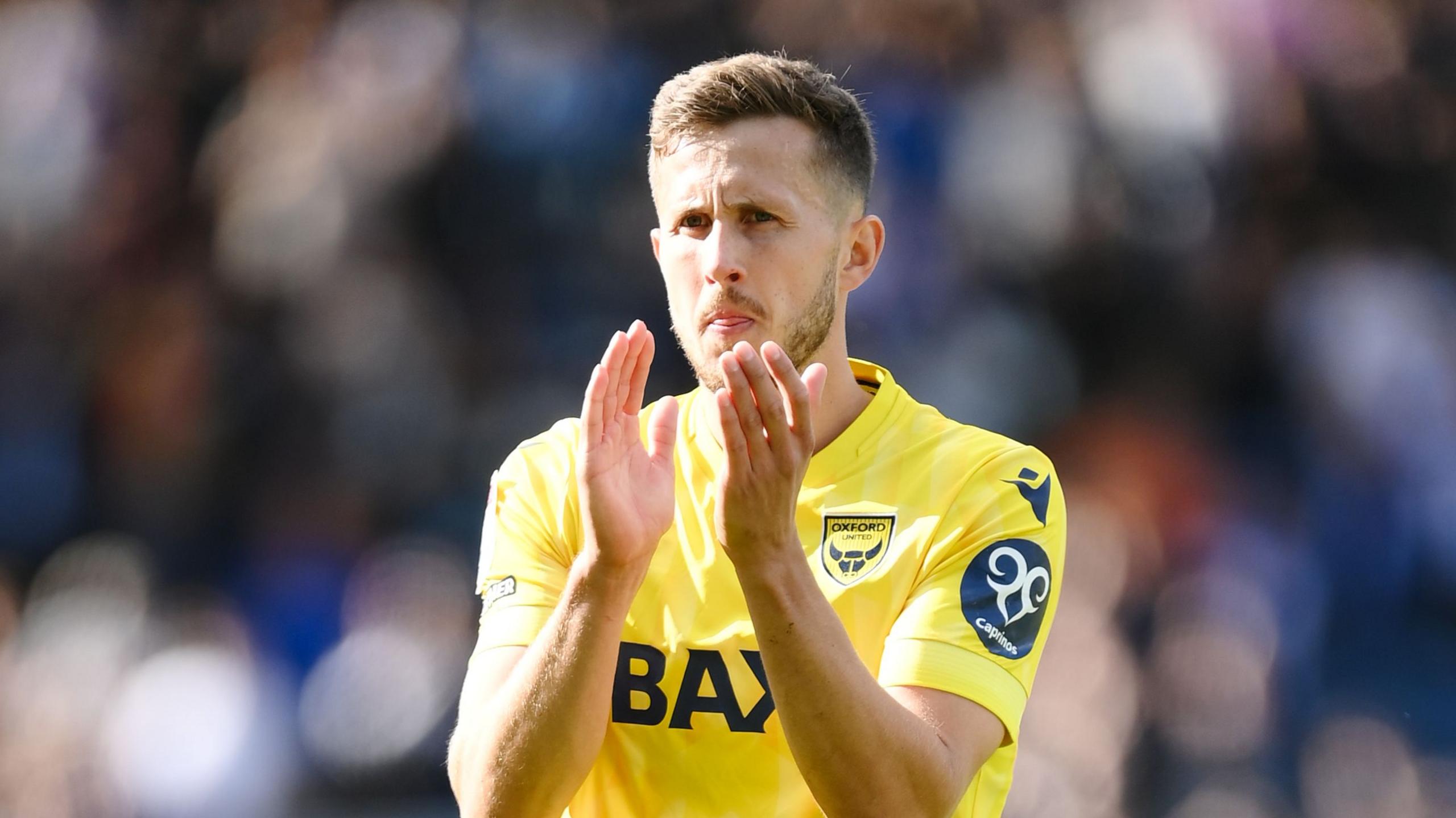 Will Vaulks of Oxford United claps following a Championship game. He is wearing a yellow Oxford kit and has short brown hair.