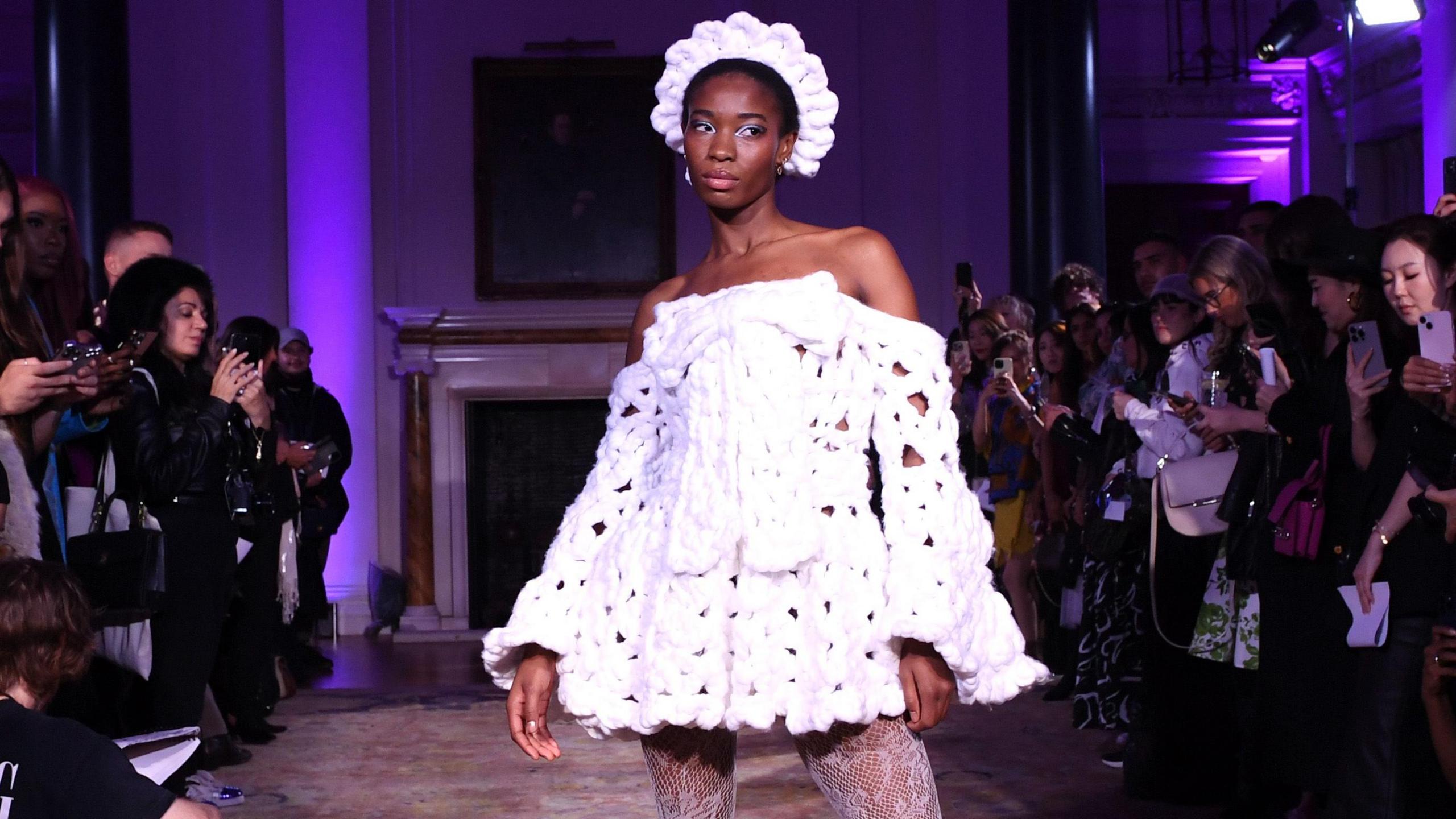 A model wearing one of Shonagh's designs at London Fashion Week. The off-the-shoulder mini dress is made from white yarn, has long flowing sleeves and is finished with a large bow on the chest. The model wears a matching crown-style headband. She poses in the purple-lit room in front of a crowd taking photos on their smartphones.