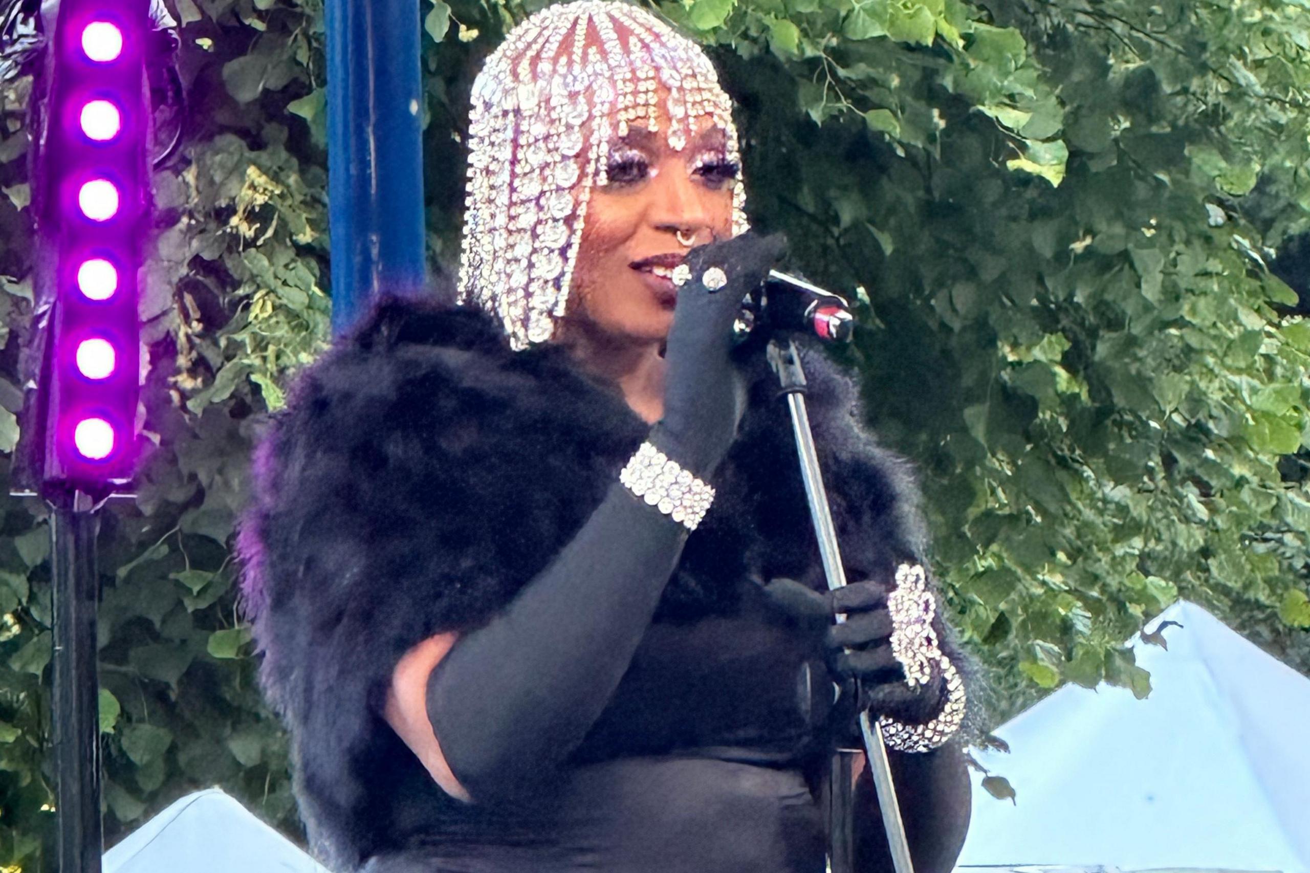 Janice Robinson sings into a microphone as she stands on the bandstand in Pageant Gardens. She is wearing a diamante-style skull cap, black gloves, a black top with a black fur-look shrug over her shoulders.