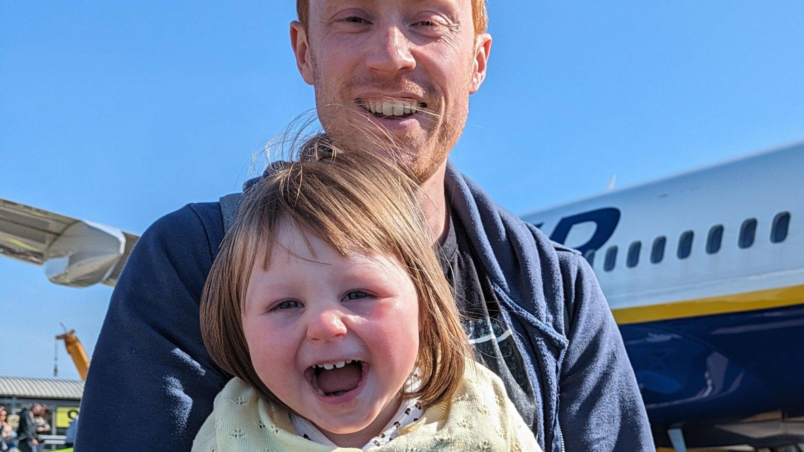 A little girl grins at the camera while her dad holds her. He has short ginger hair and she has shoulder-length brown hair. They are standing on an airport runway with a plane in the background.