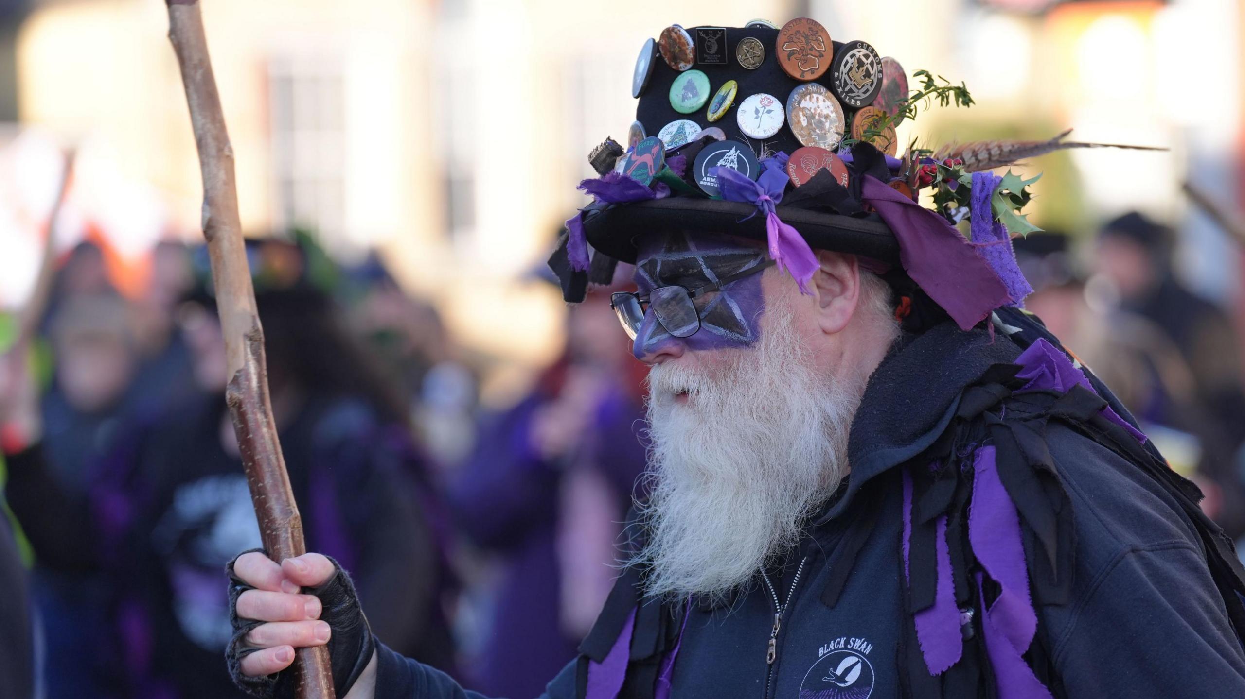 A man from the Black Swan Border Morris group is dressed in black and purple. He is not facing the camera but has a long grey beard and has black and purple face paint on.