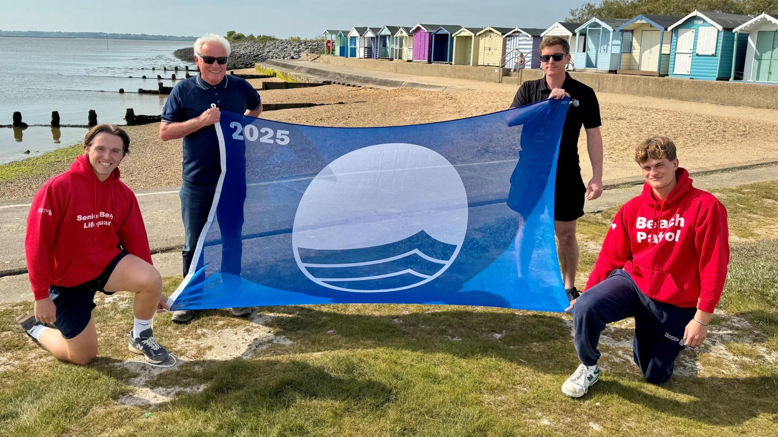 Four people holding a blue flag on Brightlingsea beach. Mick Barry, wearing navy blue and sunglasses, is second from left.  Another man in sunglasses and a dark polo shirt and shorts holds the top right of the flag. Two mean in read hoodies hold the bottom corners of the flag. Behind them is the sea, sand, groynes and beach huts.