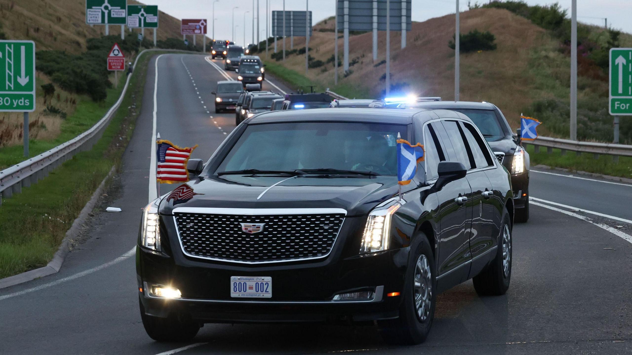 US President Donald Trump's motorcade on the A77 in Maybole, South Ayrshire, as arrives to begin his five-day private trip to the country. His black car is adorned with US and Scotland flags, and is followed by other black cars, all with their headlights on. 