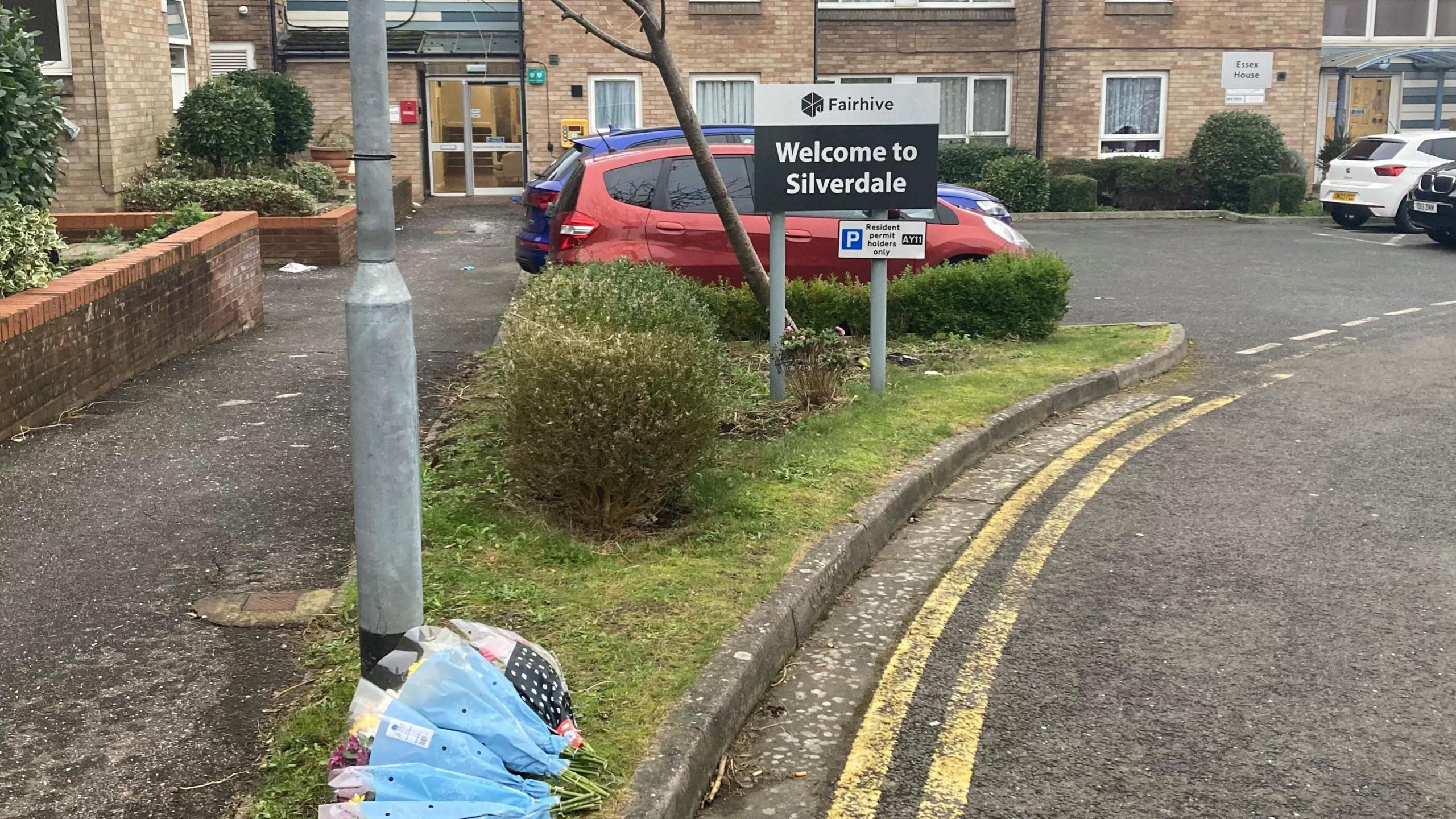 Flowers laid on the ground by a lampost in front of a small car park and blocks of flats which form sheltered accommodation. There is a sign which reads "welcome to Silverdale".