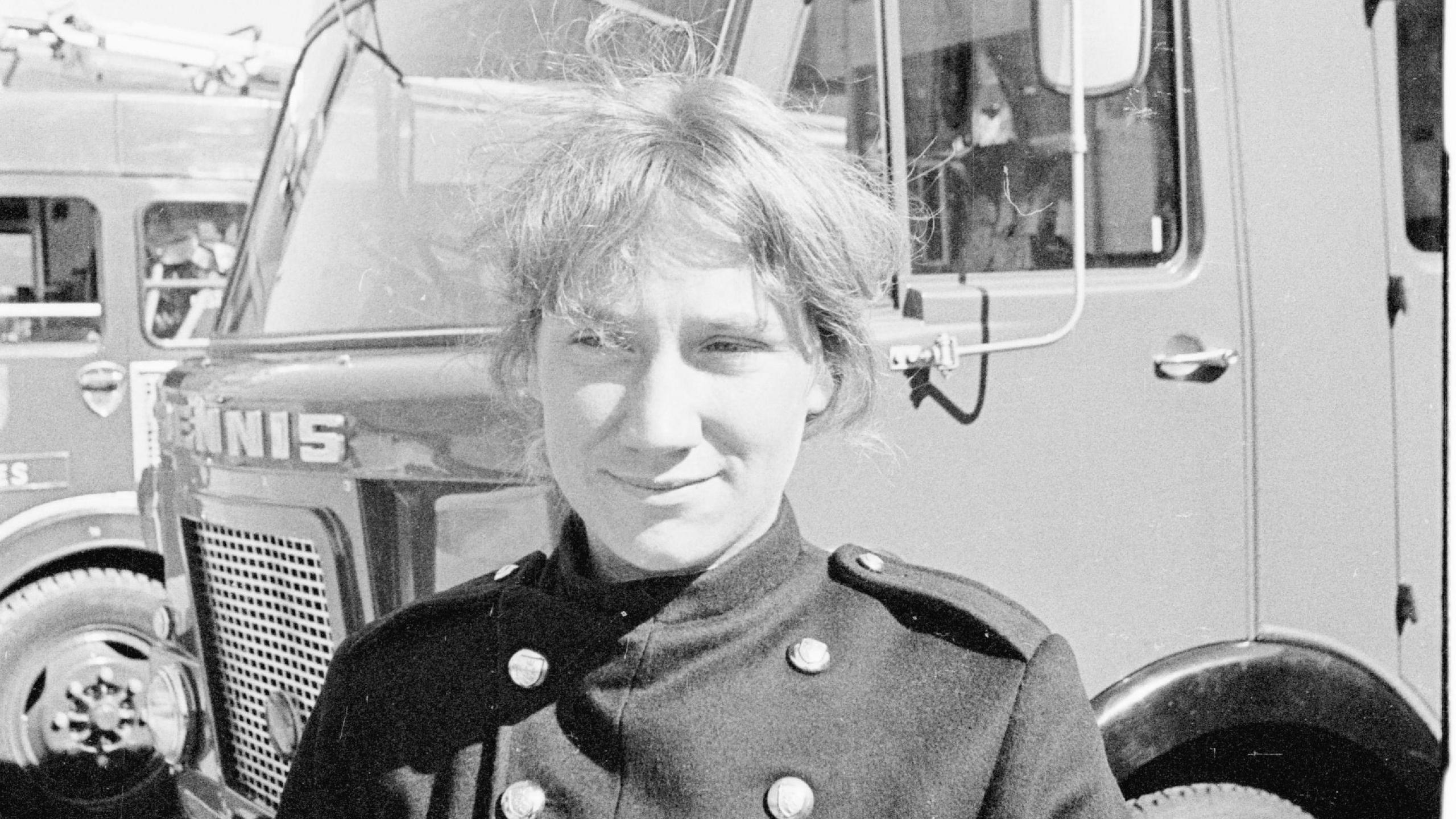A young Mary Joy Langdon in a firefighter uniform. She is stood in front of a fire engine in the black and white image.