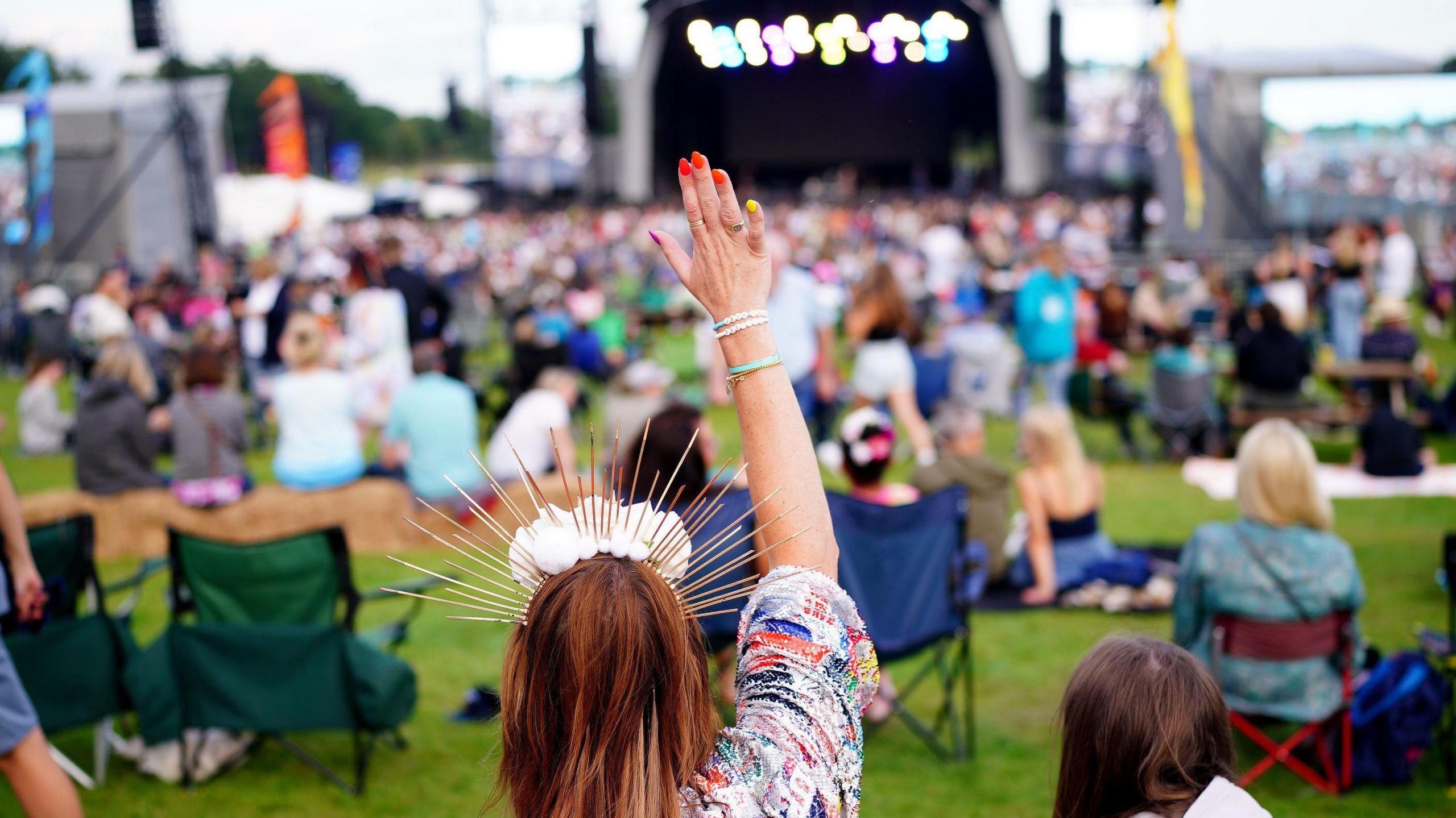 A woman with red hair and a sparkly headband holding her hand up, with a stage and crowd out of focus in the background