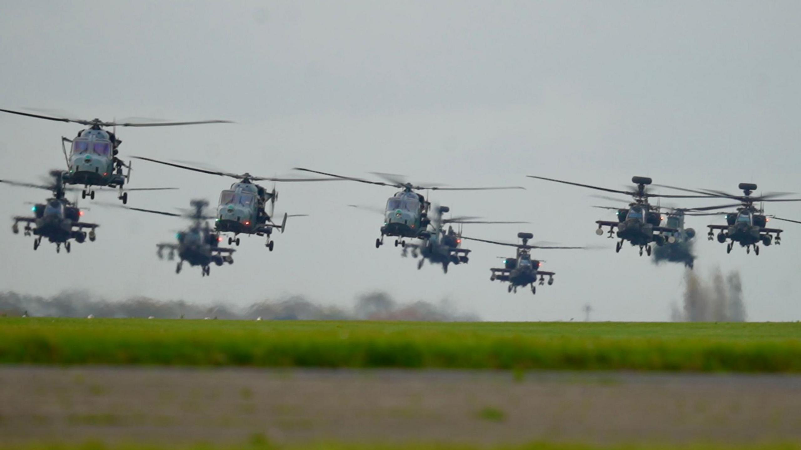 Dozens of helicopters lift off from an airfield on a grey day. They are in a close formation as they fly.