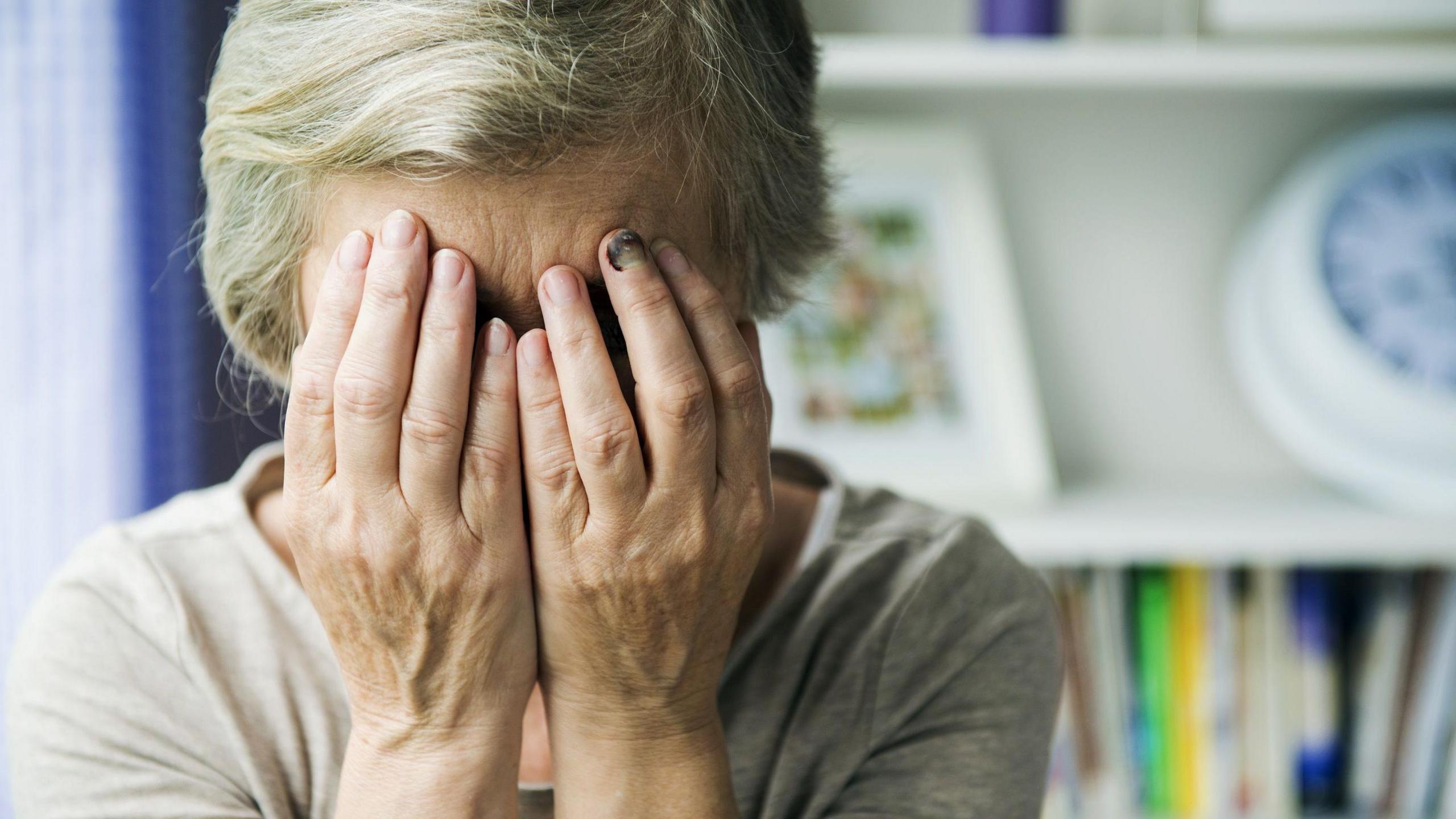 An elderly woman with white hair holds her face in her hands