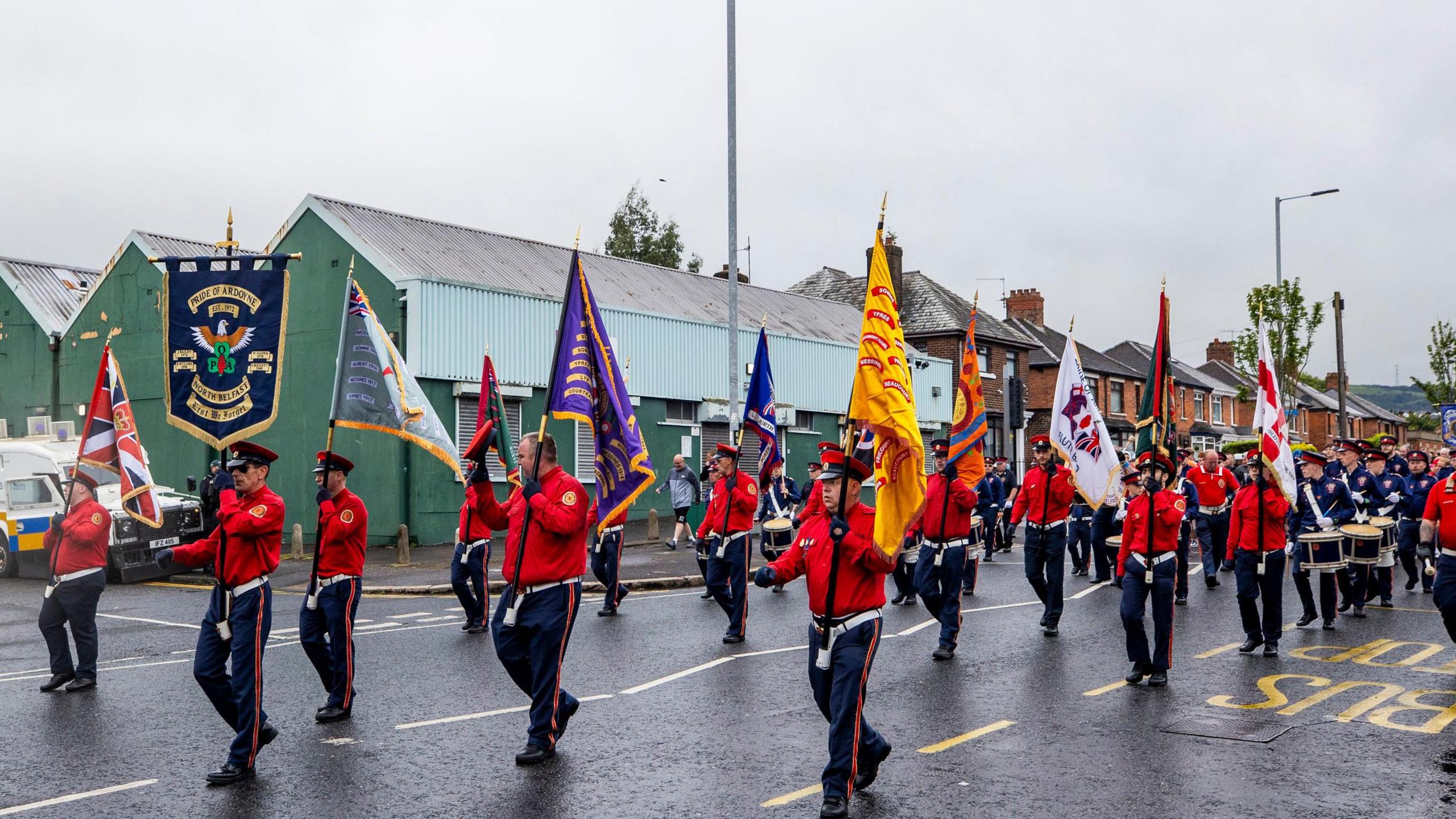 Ranks of band men wearing navy trousers and red shirts, holding flags, lead ranks of drummers and flautists on parade.