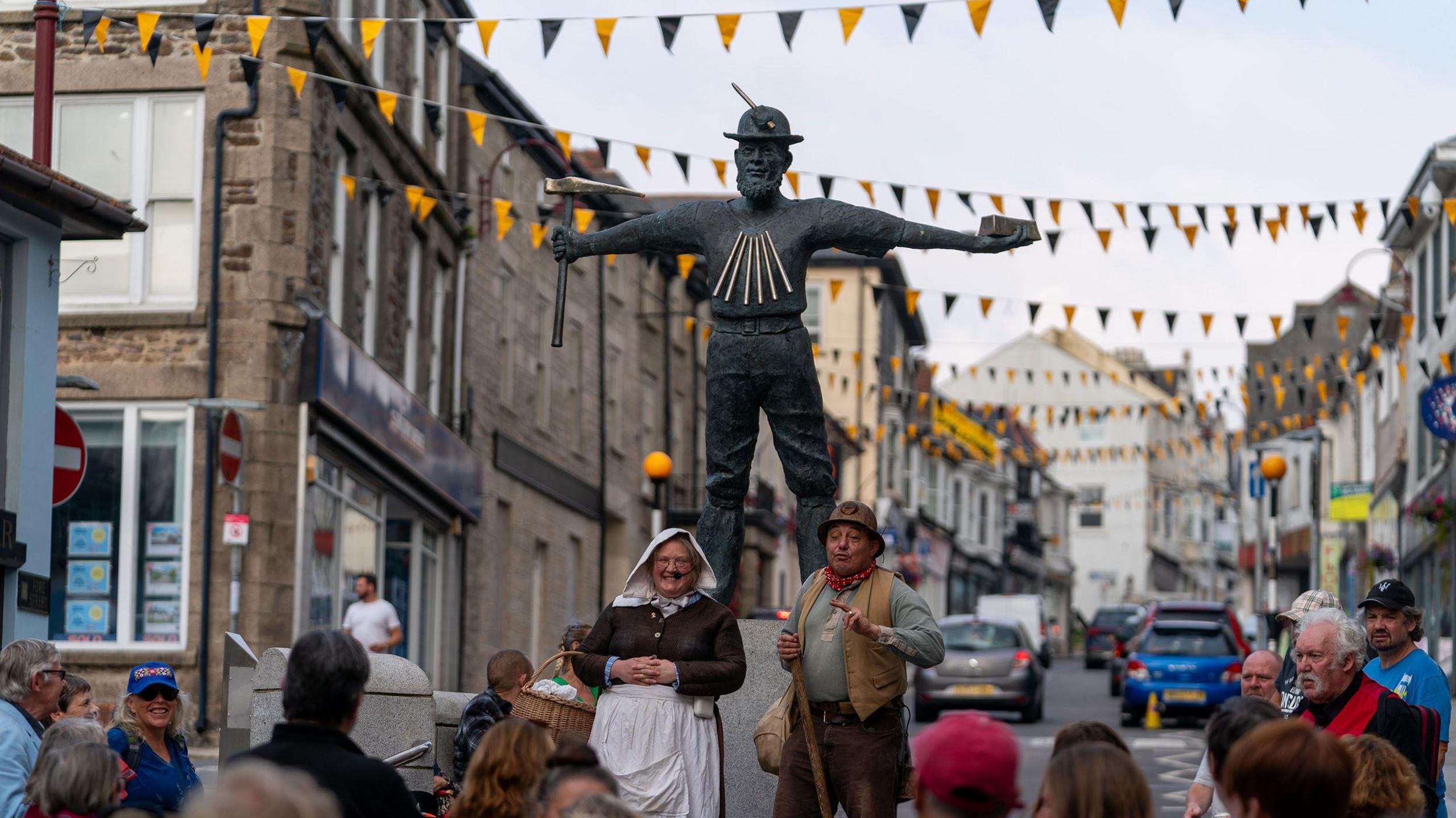 There is a man and a woman in period dress standing under the Tin Miner Statue in Redruth surrounded by a crowd of spectators. The statue is grey and shows a miner wearing a helmet with his arms outstretched holding a pickaxe and a rectangle-shaped ingot. She is wearing a white hood and apron and a brown dress and is holding a basket. He is wearing a brown waistcoat and hat with a red and white spotted neckerchief over a green short with brown trousers. They are both smiling as they face the crowd. There is yellow and black bunting strung high over the street behind them. 