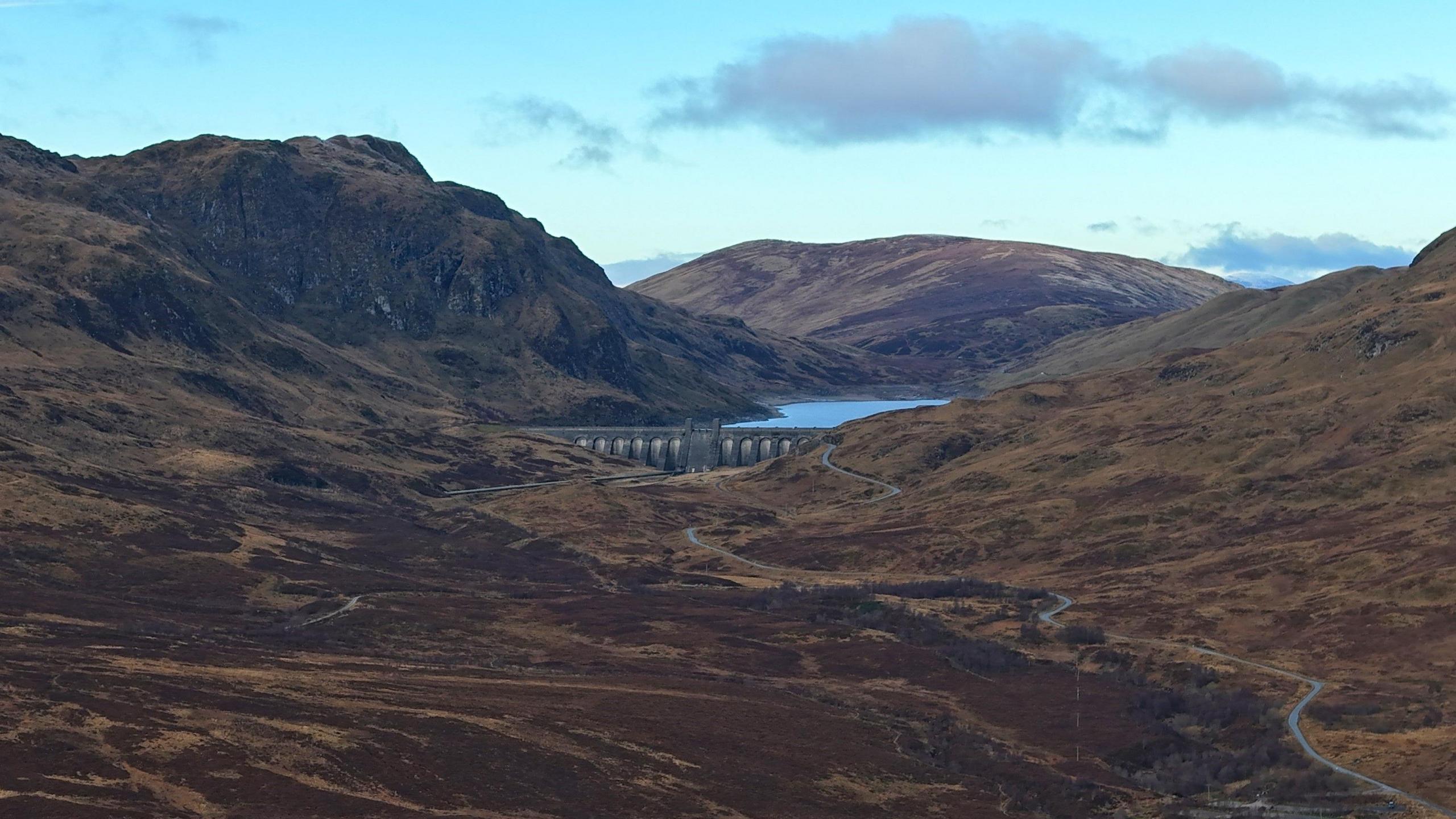 The dam is in the middle of the picture and you can just make out a strip of water behind it. It is surrounded by dark brown hills and a blue sky.