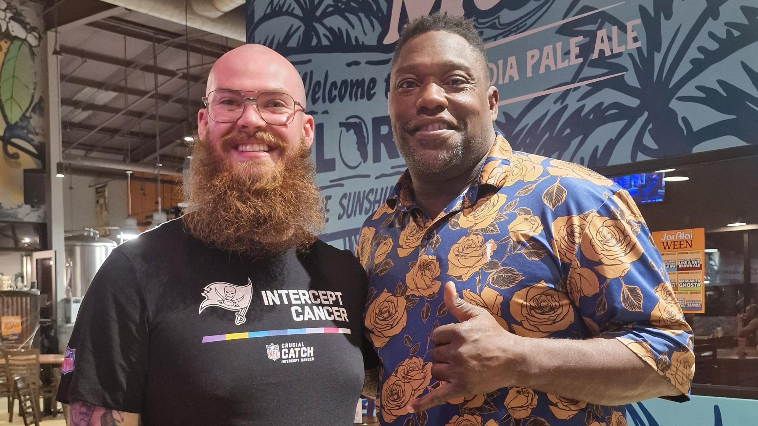 Josh stood with former NFL player Warren Sapp. Both are smiling into the camera. Josh is wearing a black Bucs t-shirt while Warren is wearing a blue and orange floral shirt.