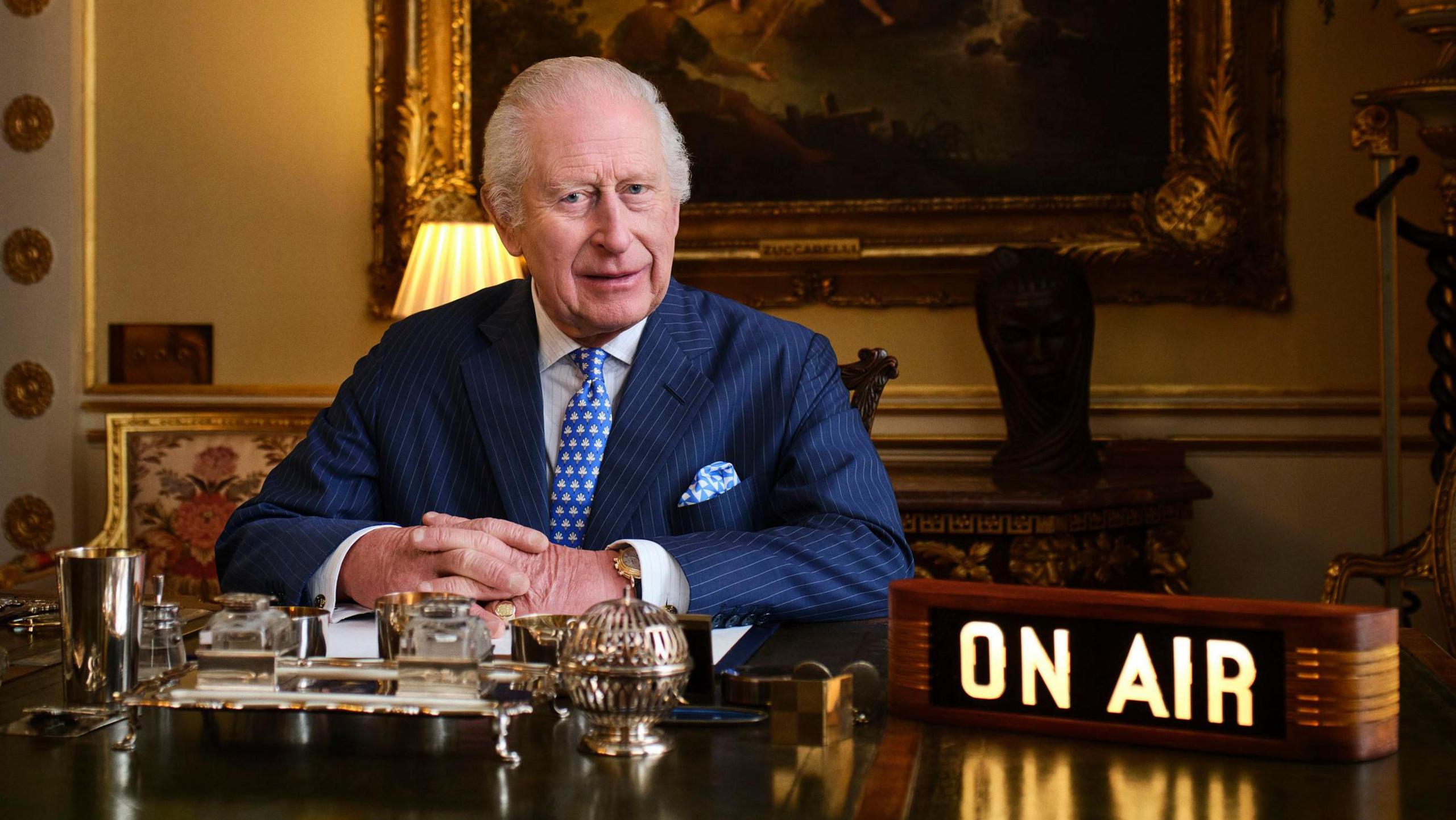 King Charles sitting behind a desk with an 'on air' sign beside him