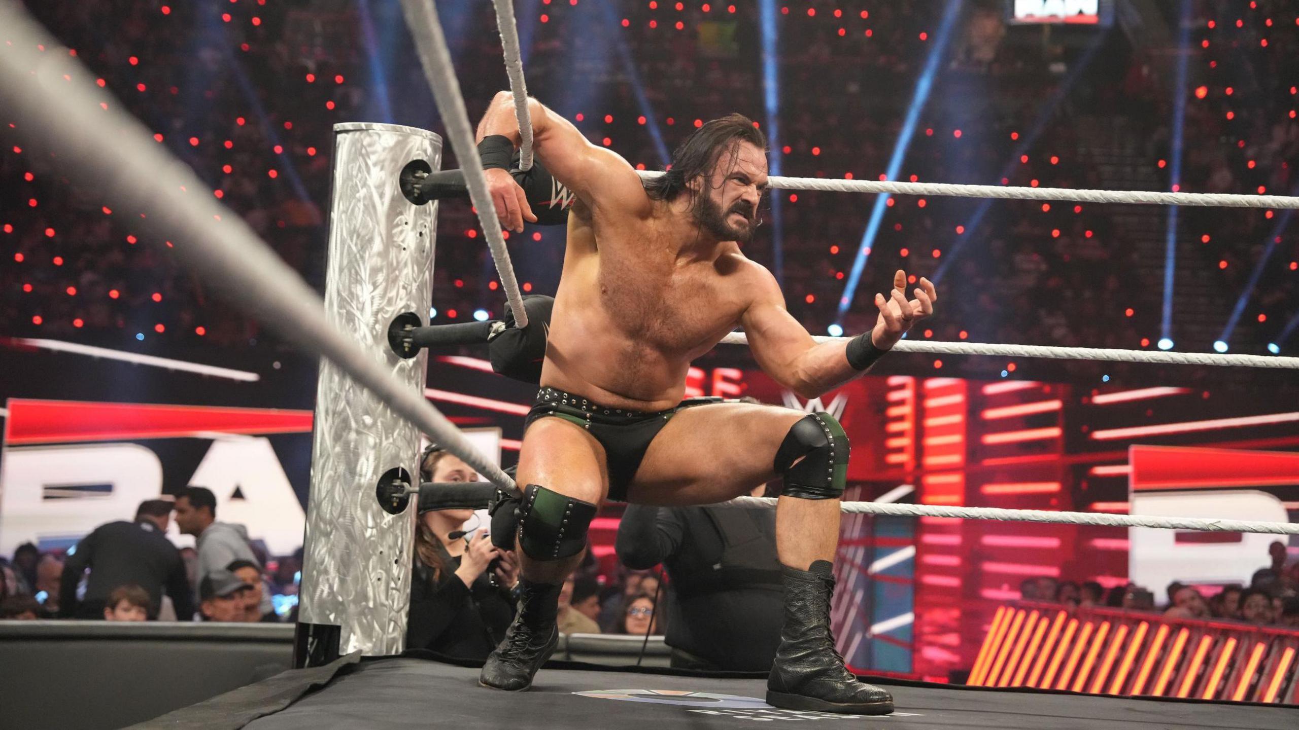 Drew McIntyre in the ring, crouching by the turnbuckles, with one arm resting on the ropes. His face is in a grimace, while he is wearing black trunks and boots. He appears about ready to pounce on an opponent. 