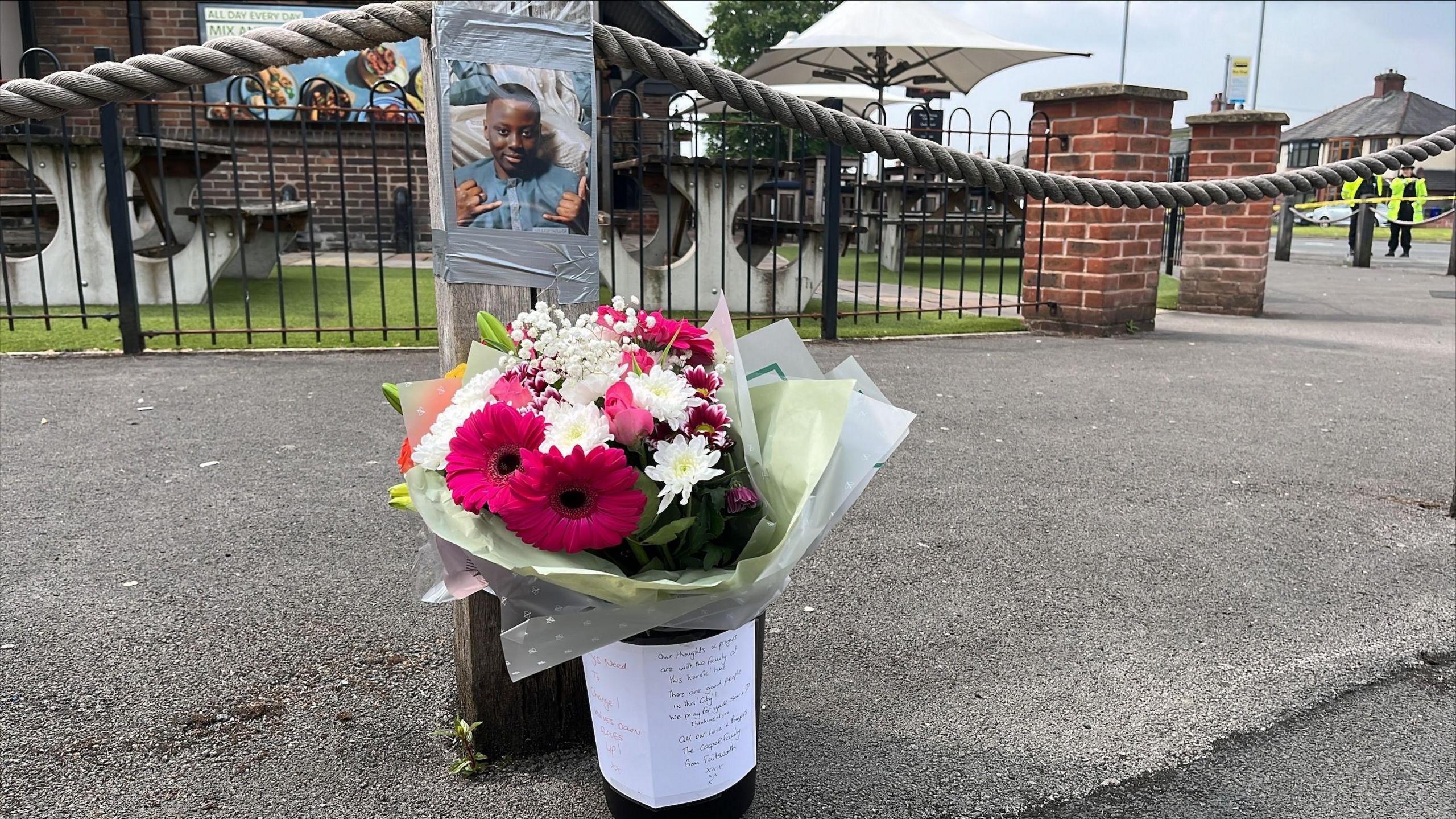 A bucket of flowers with tributes attached on paper, underneath a wooden post with an image of Ibrahima attached. The tributes have been left on a street.