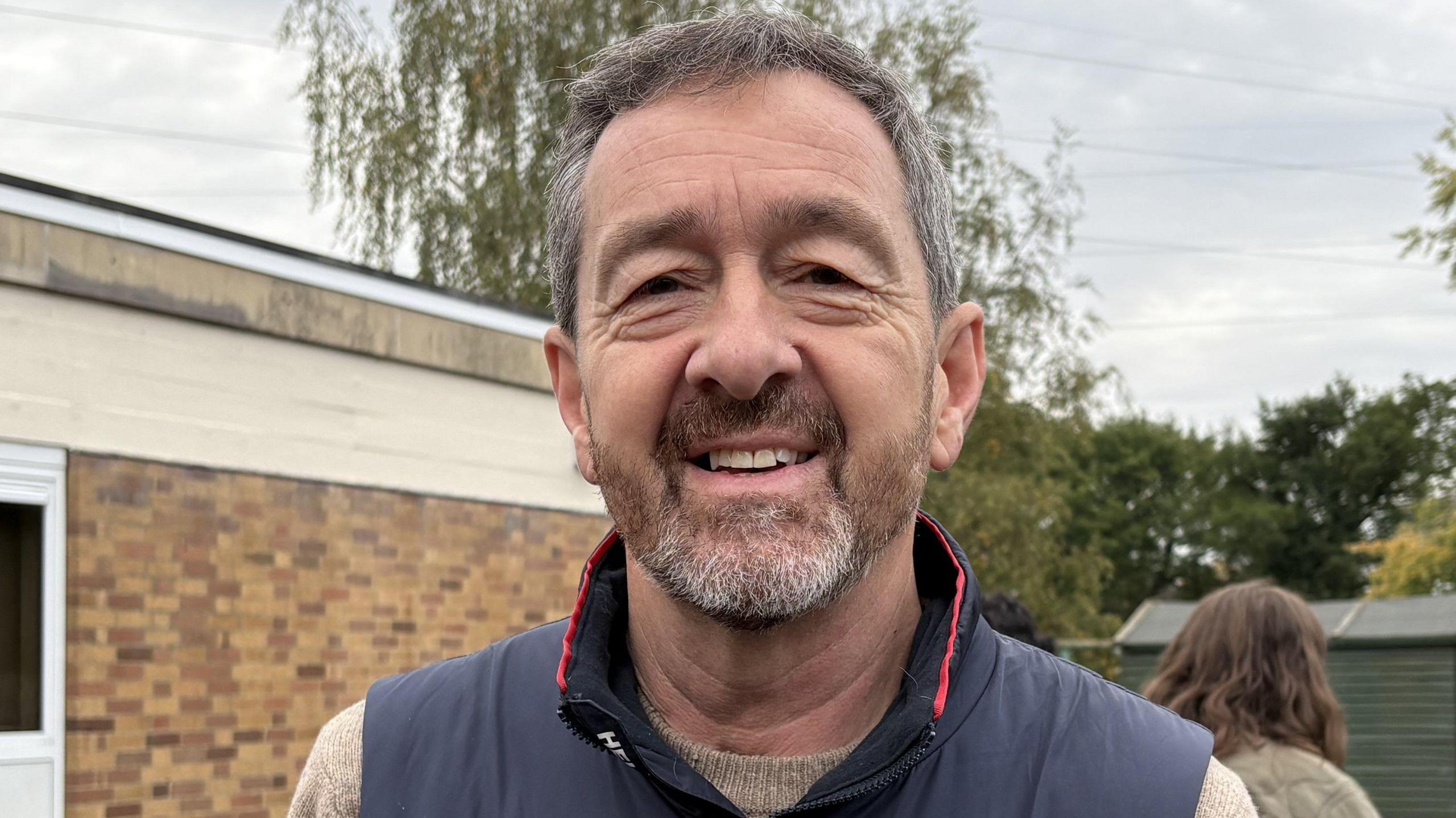 A man with grey hair and facial hair smiling at the camera. He is wearing a navy blue gilet and beige jumper underneath