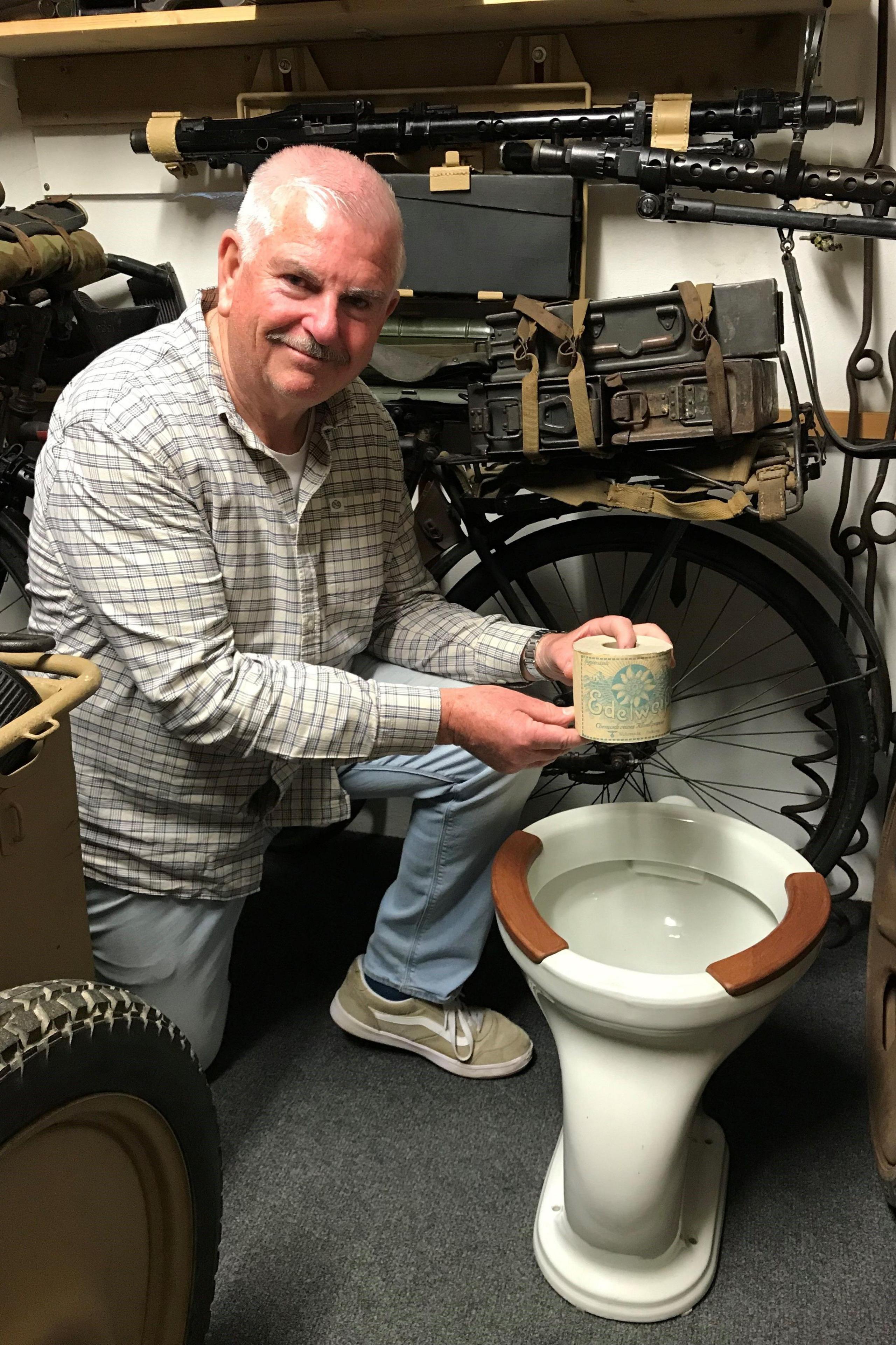 Graeme Delanoe kneels holding a roll of occupation-era toilet roll above a toilet bowl with is white porcelain with two brown wood panels around the top of the bowl. Behind are various WW2 items stacked on shelves.