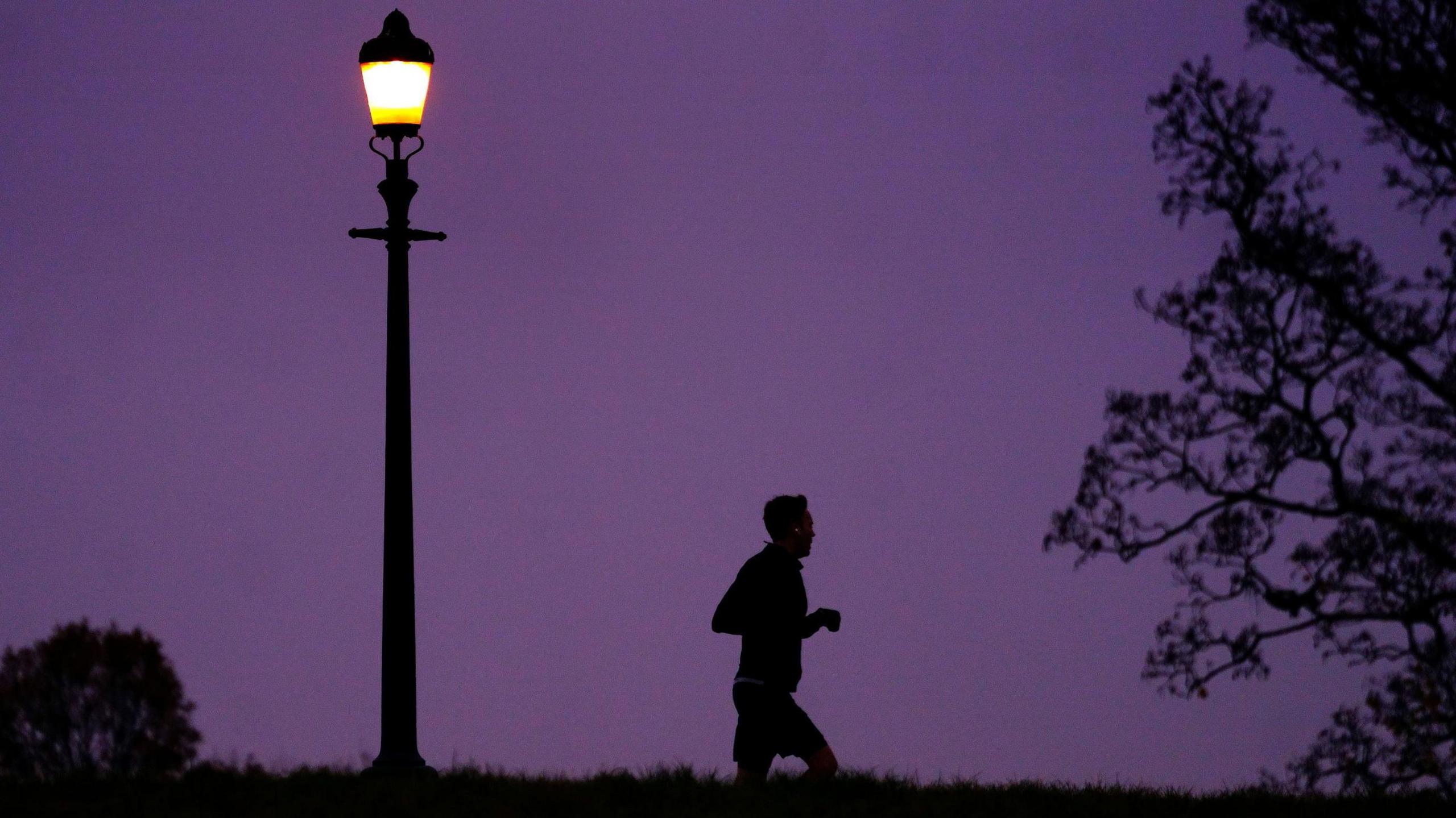 A silhouette of a man dressed in shorts can be seen jogging in a field. He is running towards trees and there is a street lamp to his left and there is a purple night sky behind him.