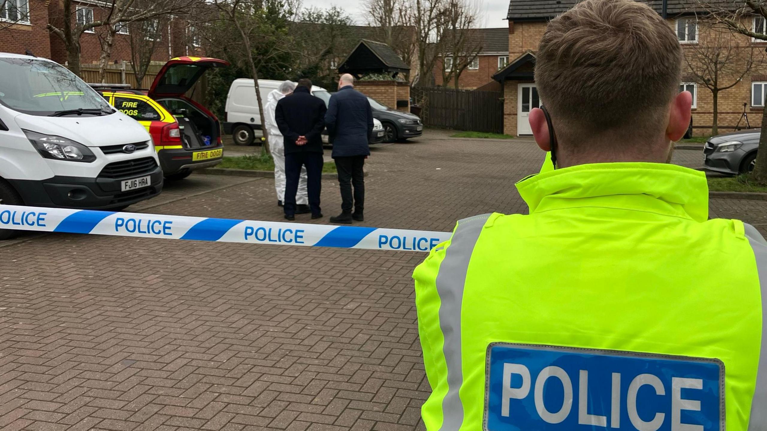 A police officer stood at a police cordon. Detectives and forensic officers are stood inside the cordon. 