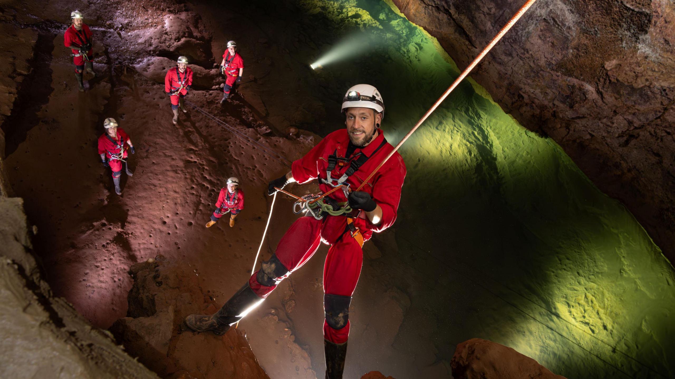 A caver in red overalls and a white safety helmet is suspended by rope over a large cavern at Wookey Hole in Somerset. Below him are other cavers standing on the cavern floor in the same outfits.