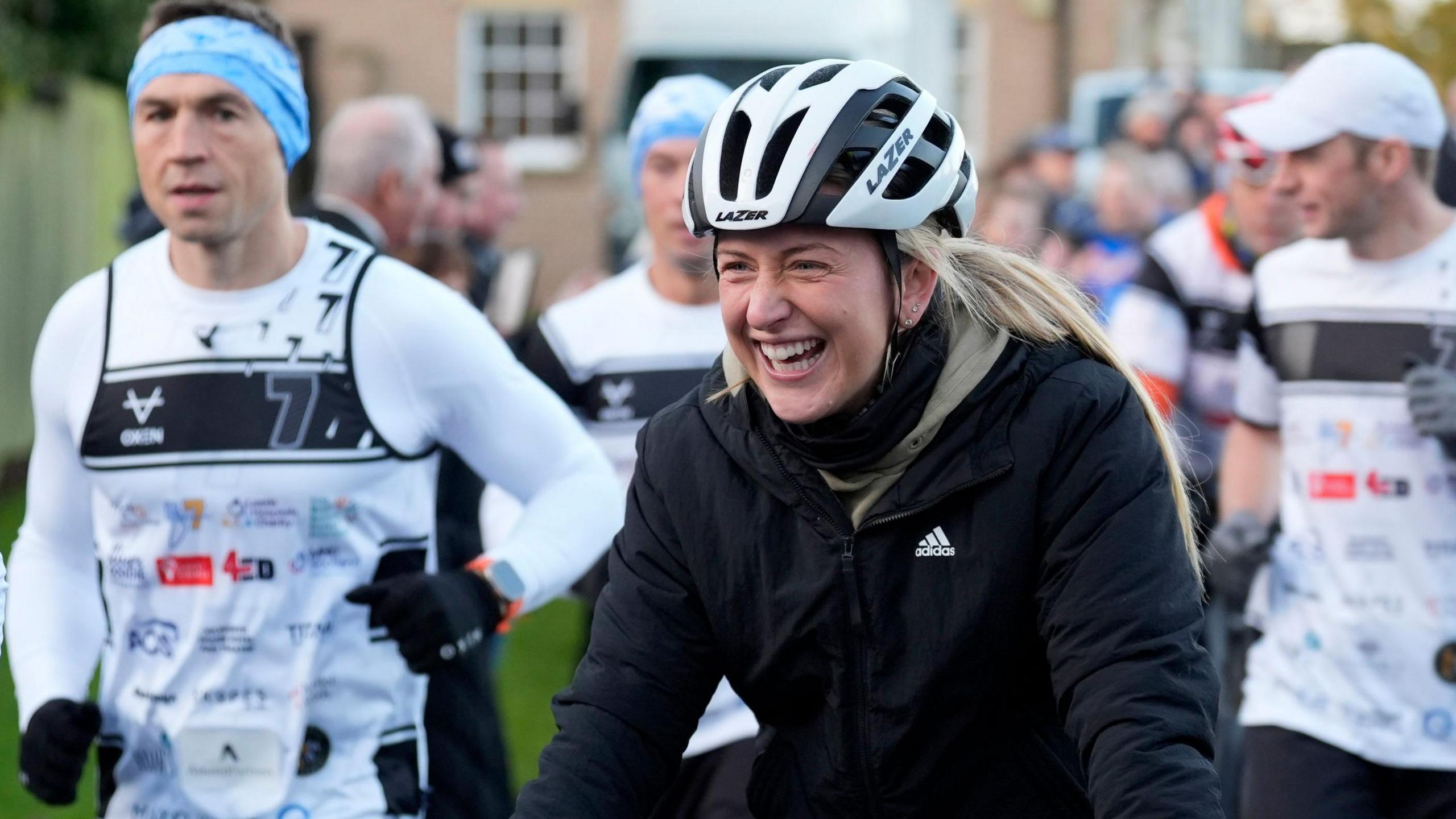 A smiling Dame Laura Kenny with a white helmet riding a bike. Sinfield and other runners can be seen in the background.