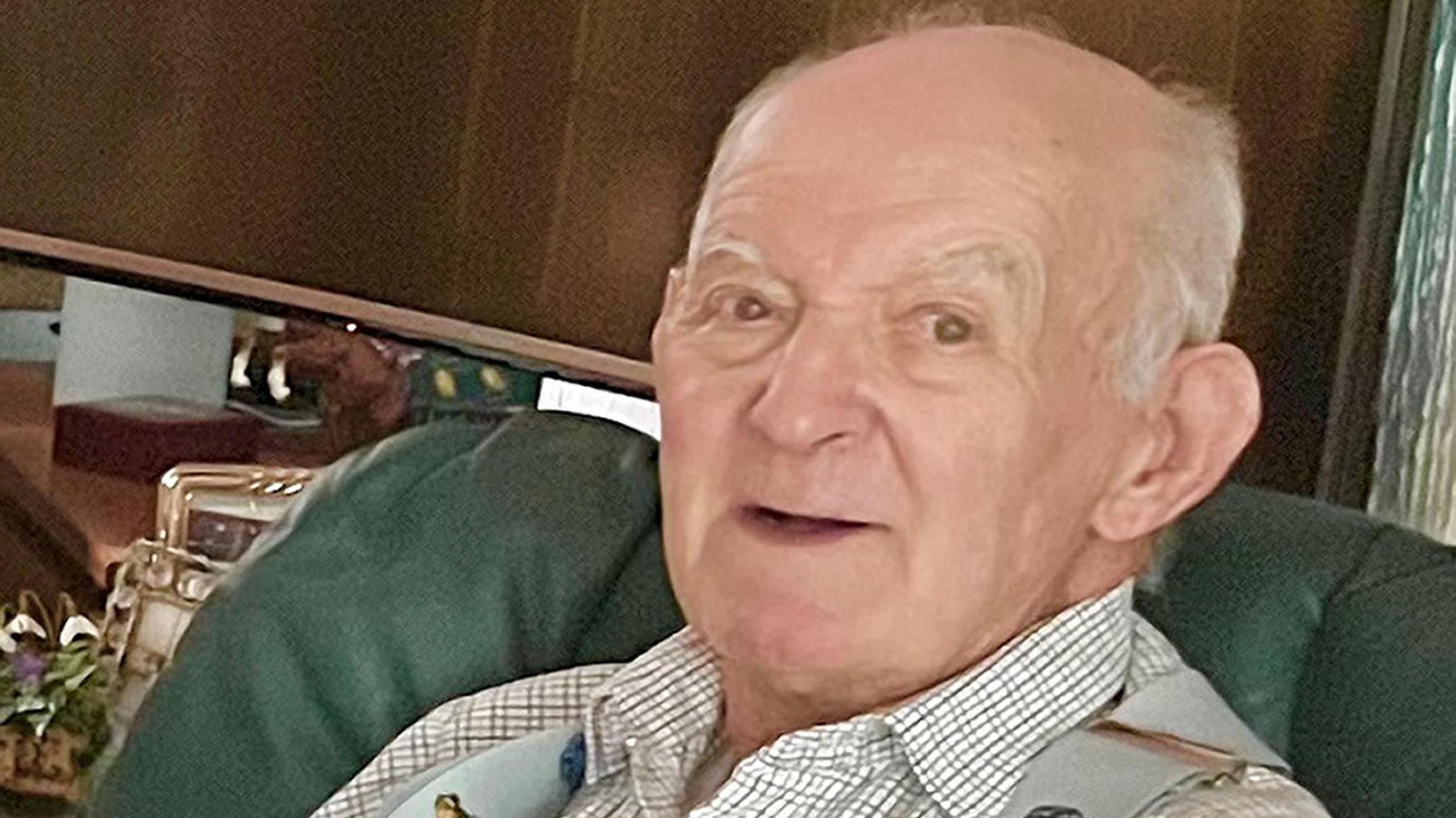 Photo of Paul Swales, an elderly man wearing a white cross-hatched shirt, sitting on a green chair and smiling at the camera