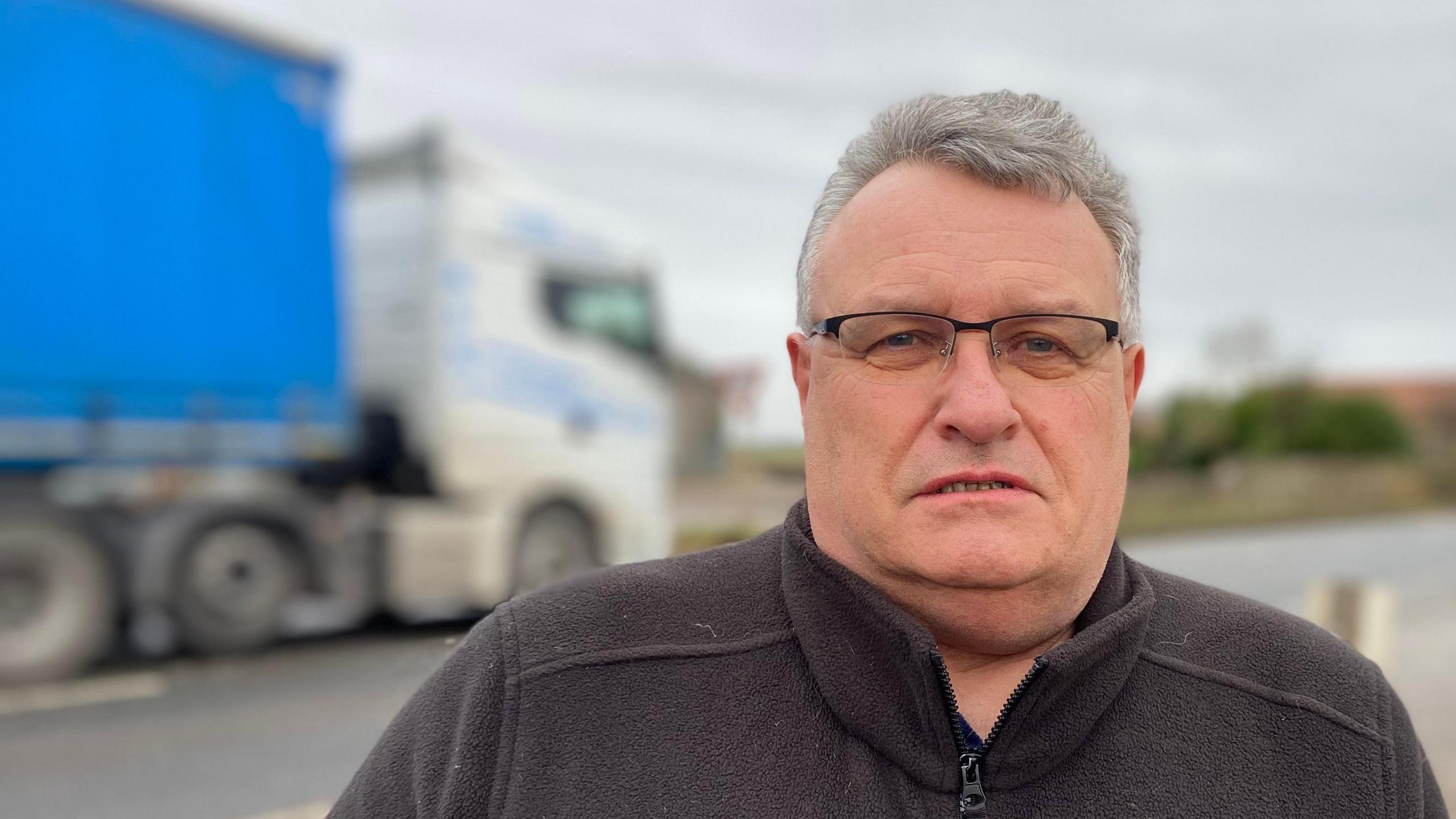 Man standing in a black fleece with a lorry passing behind him