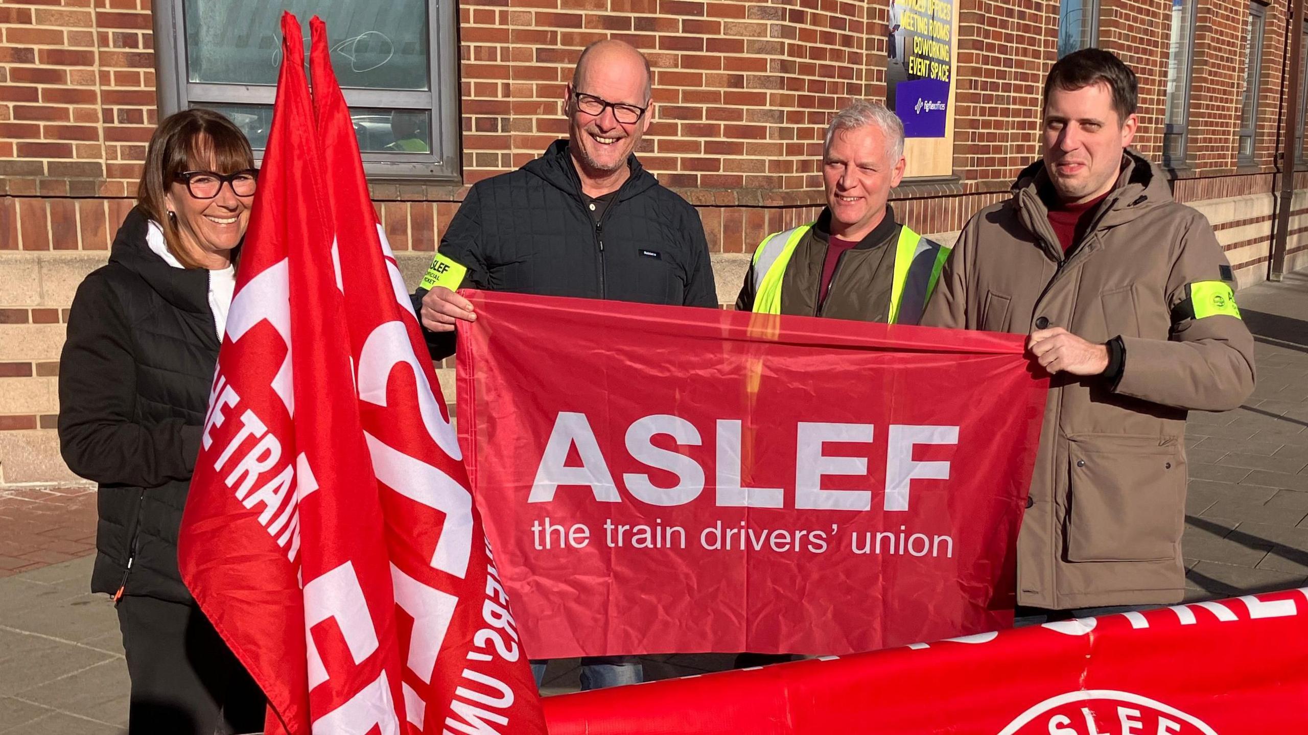 Four people stand on a street corner holding red banners and flags bearing the words: Aslef the train drivers' union. The three men and a woman are smiling and standing in front of a large red brick building.