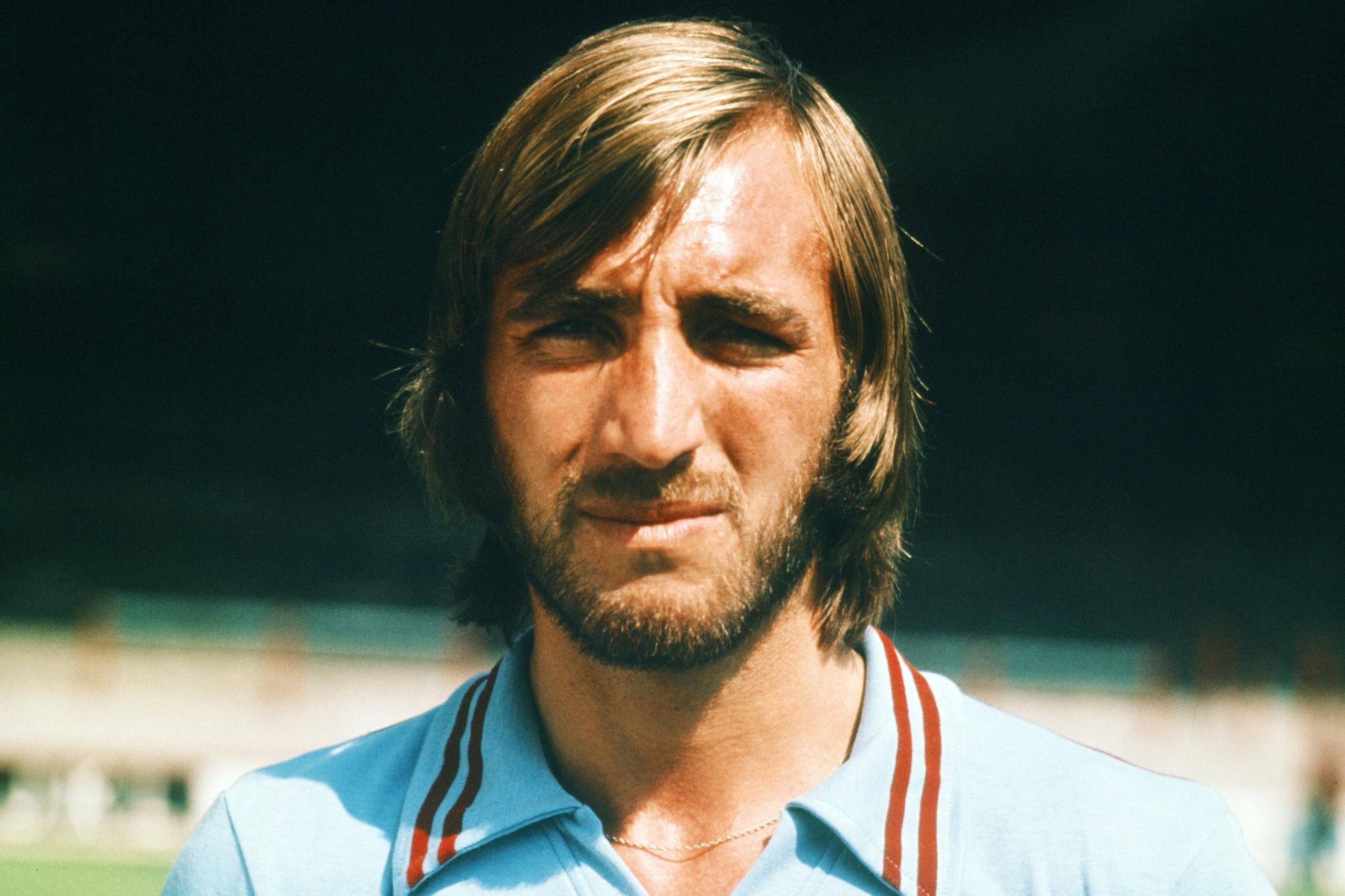 Billy Bonds ka veshur një këmishë blu të çelët me vija të kuqe dhe të bardha në jakë, duke qëndruar jashtë përpara një stadiumi.