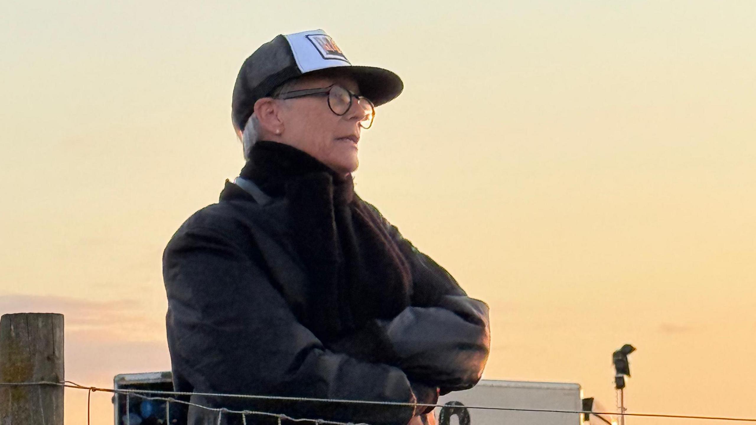 Jamie Lee Curtis behind a fence, wearing black jacket and cap