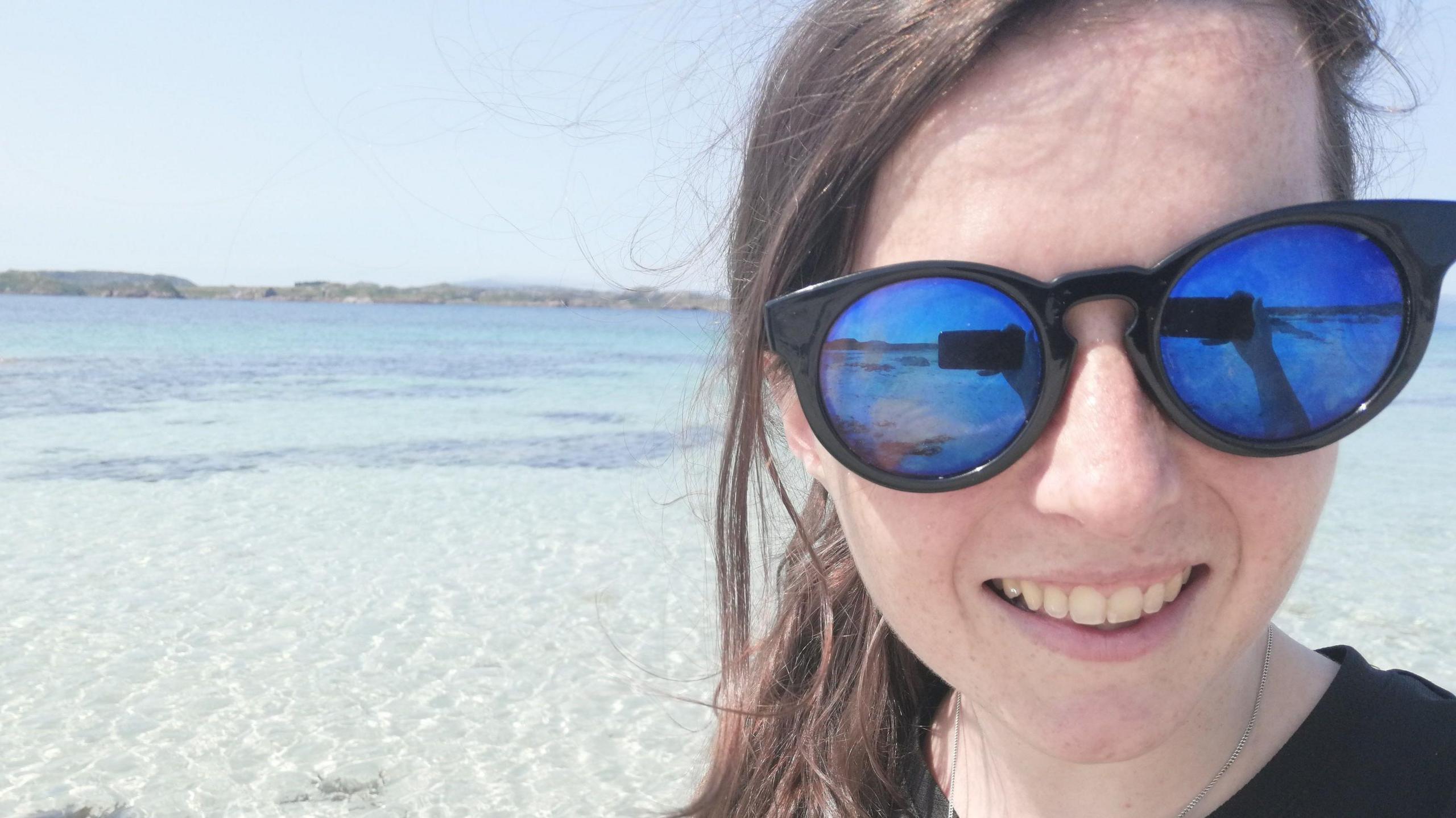 A selfie taken by Rebecca Crowe. She is wearing sunglasses and behind her is some beautiful clear water.