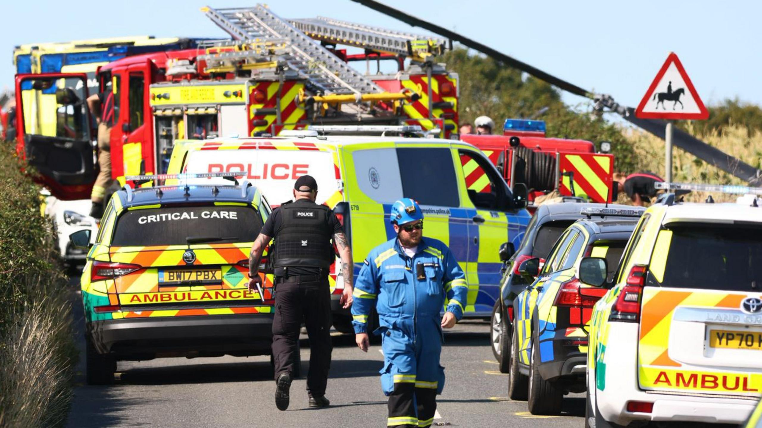 Emergency services including police officers and a man in all blue overalls, walking on a road with emergency service vehicles parked along it