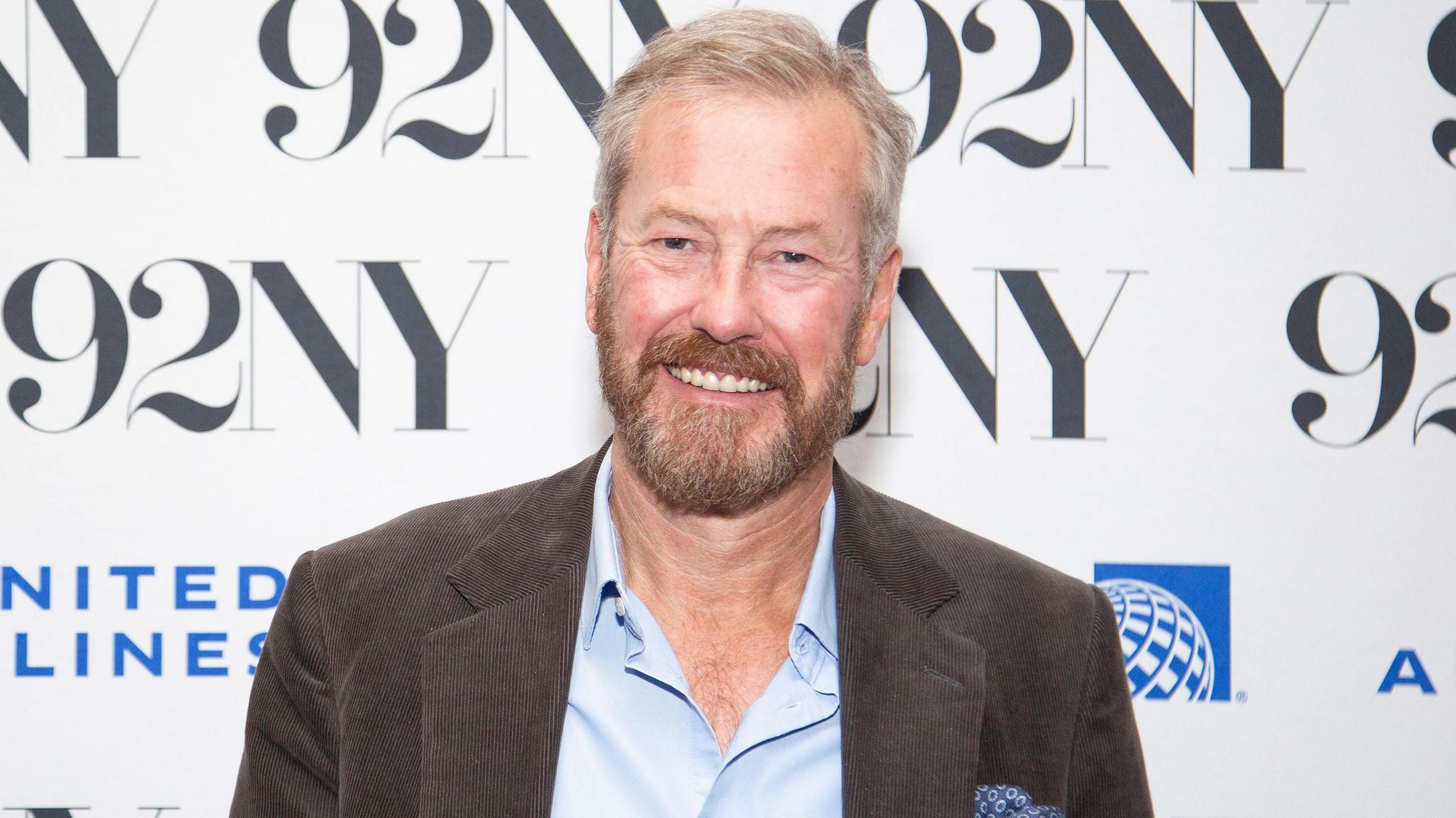 An image of a man in his 60s with short grey hair and a brown beard at a red carpet event. The event's branding, 92NY, is visible in the background. He is wearing a brown jacket, blue pocket square and a blue shirt open at the neck.