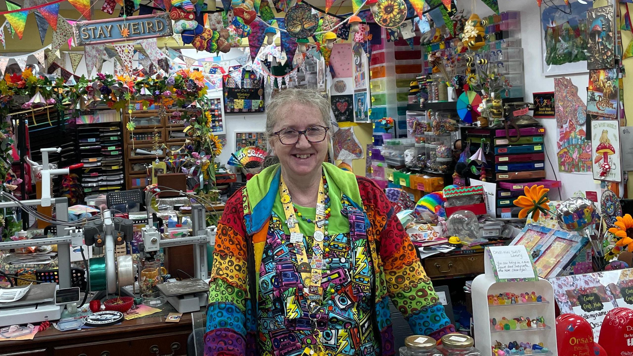 A woman wearing a multi-coloured zip-up hoodie and a very colourful t shirt stood in the opening of a shop.