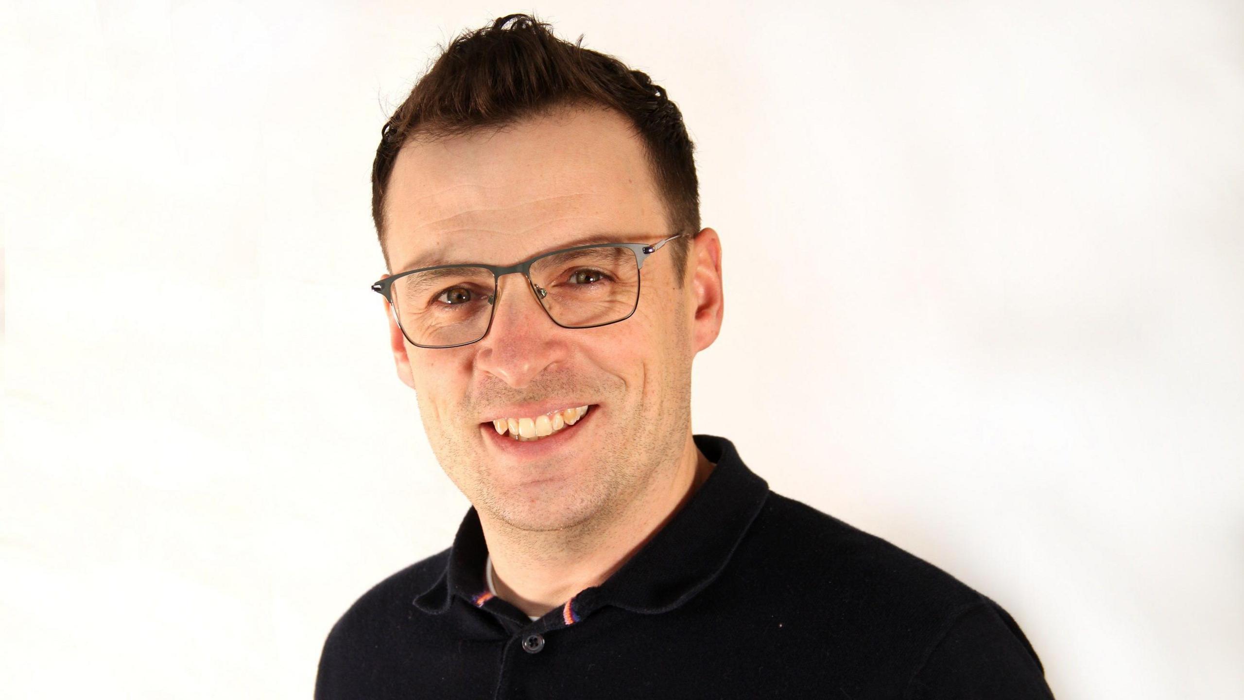 A head-and-shoulders photo of a man with short brown hair and glasses, and wearing a black polo shirt. He's smiling at the camera.