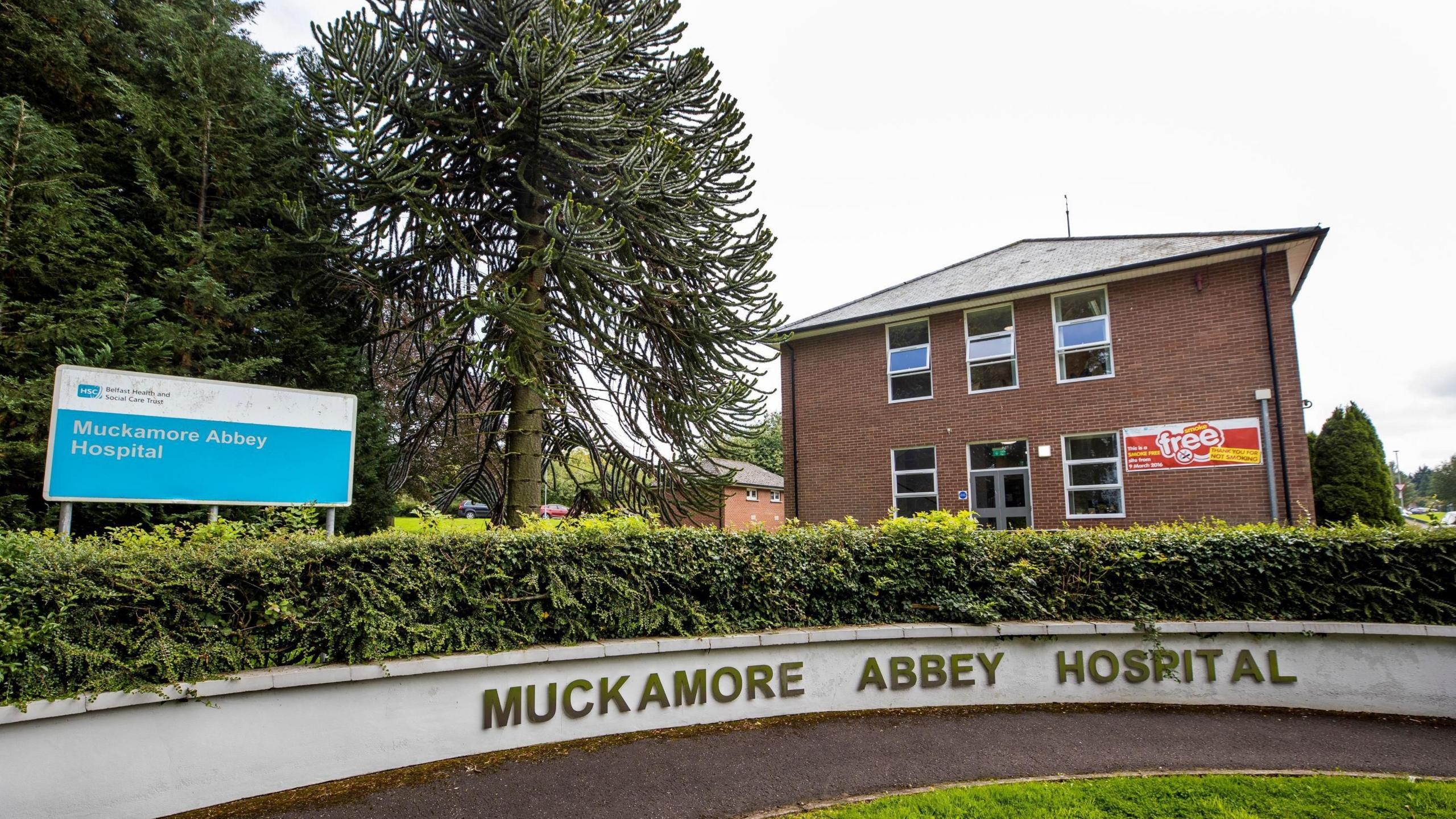 A white wall reads 'Muckamore Abbey'. There's a red brick building behind the low wall and to the left of it are green trees and a sign for the hospital.
