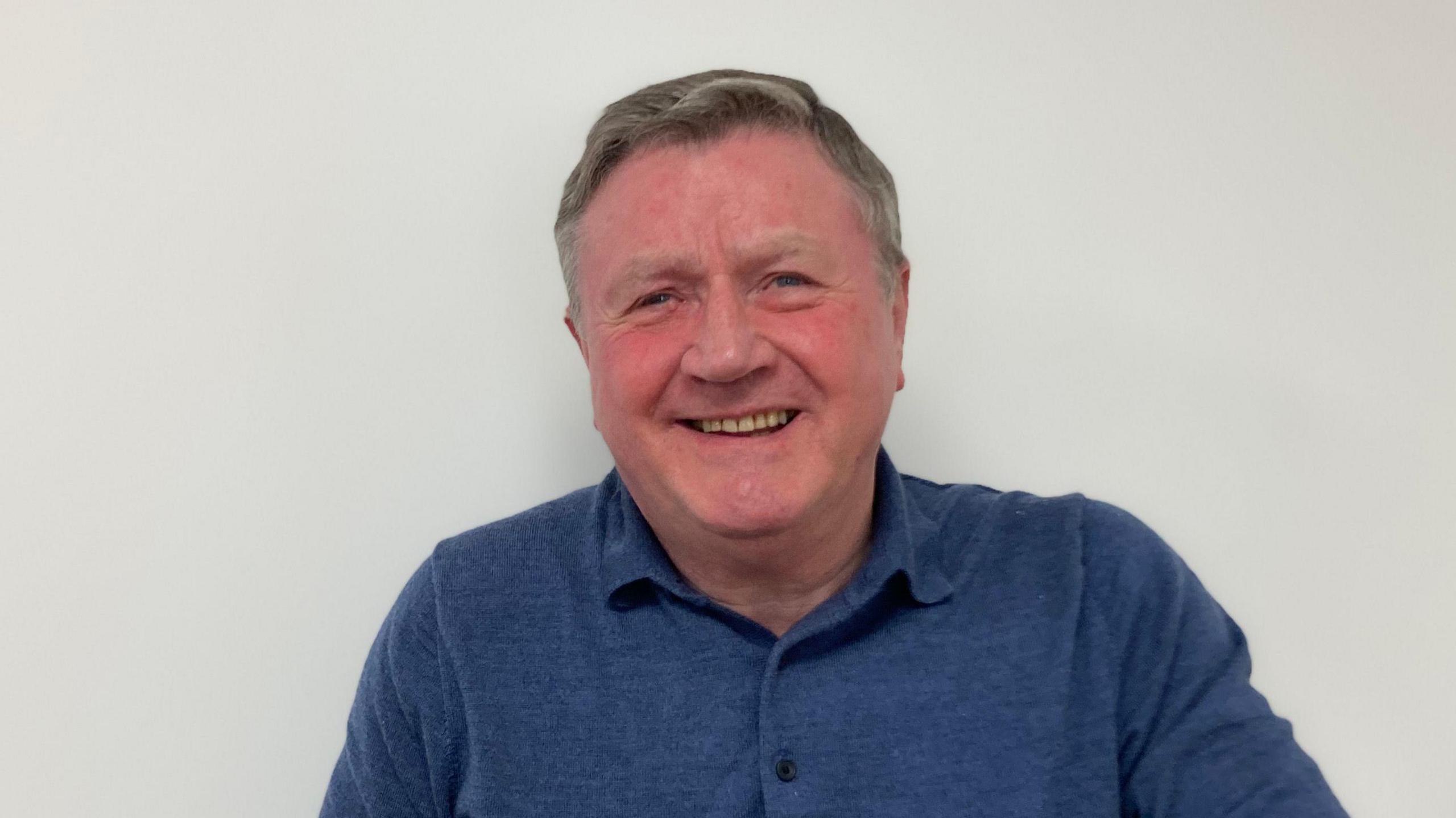 Headshot of Dennis Jones smiling at the camera against a white wall background, with a blue polo shirt