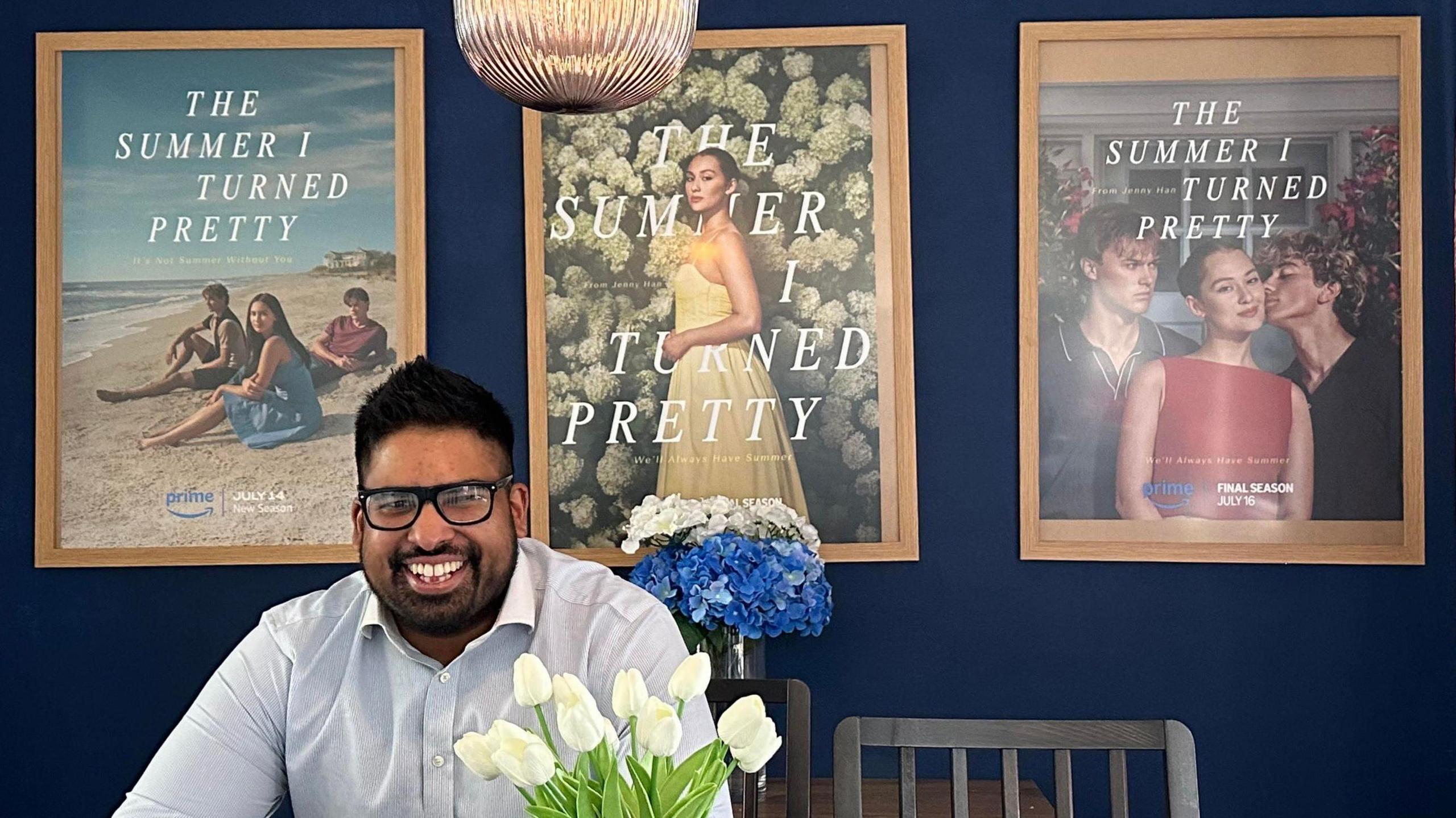 A man wearing glasses with dark short spiky hair, smiles at the camera as he is sat on his dining chair. Behind him are three posters from the series The Summer I Turned Pretty 