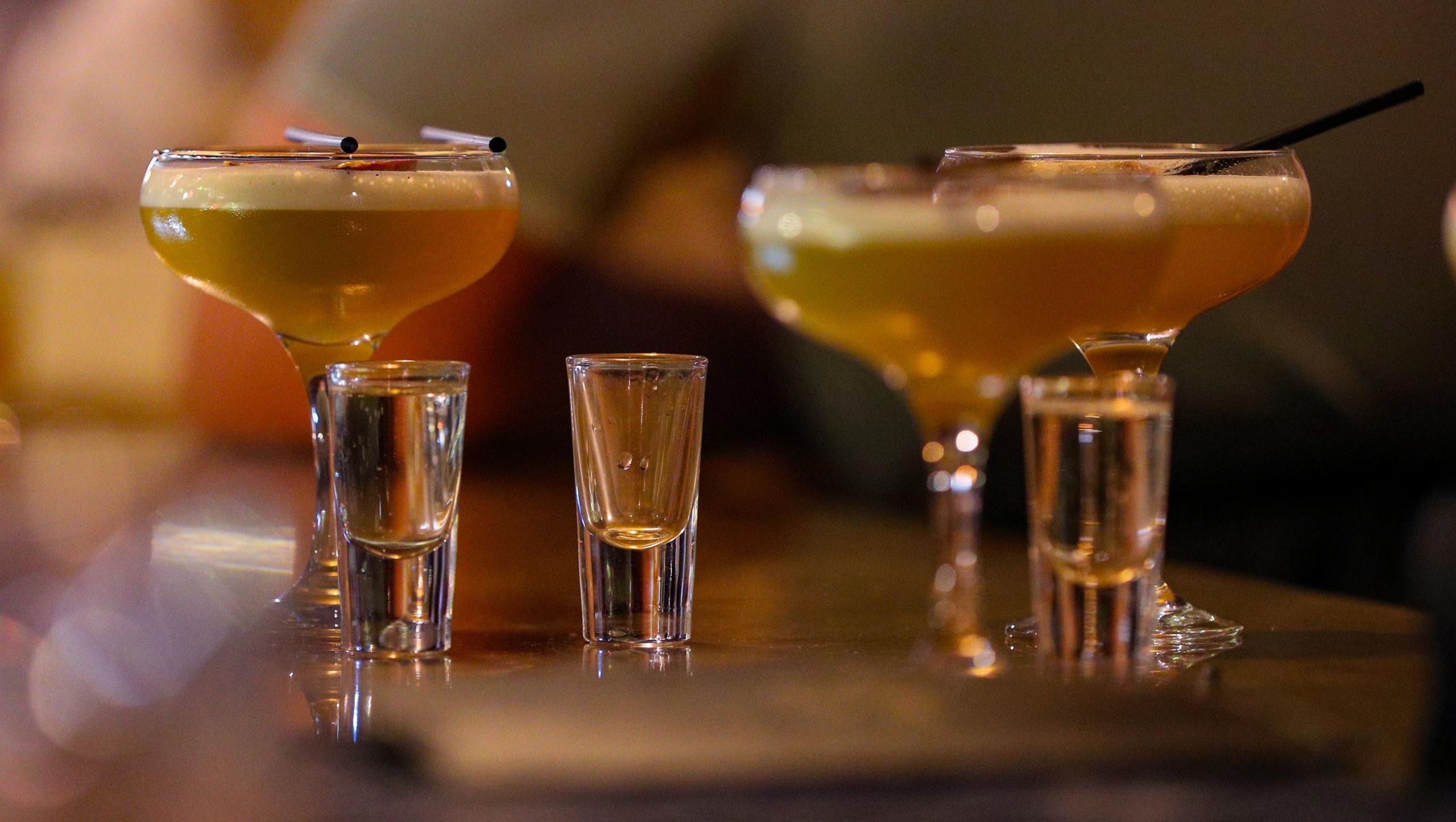 Three cocktails of amber colour with a foam top and three shot glasses filled with a clear liquid on a bar.