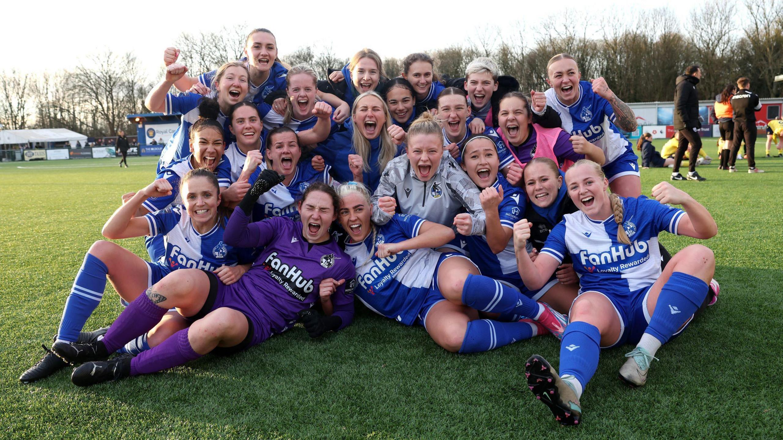 Players of Bristol Rovers pose for a group photo on the pitch as they celebrate victory after the Adobe Women's FA Cup Third Round match against Oxford United. They are smiling and laughing and many of them are punching the air. The players are wearing the traditional Rovers kit of blue and white quarters.