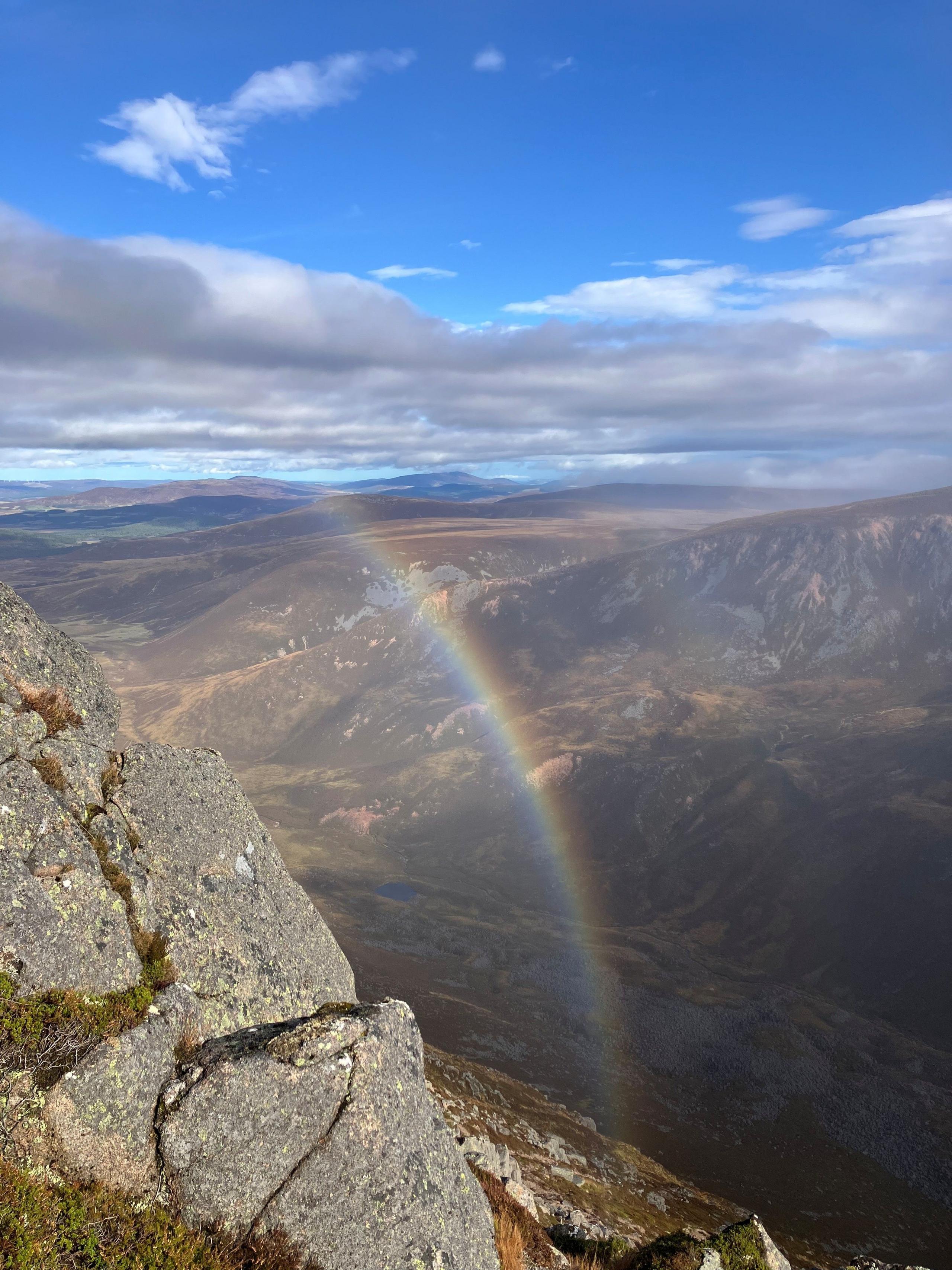 Bright blue sky with a rainbow going into a valley between two mountains in the Cairngorms