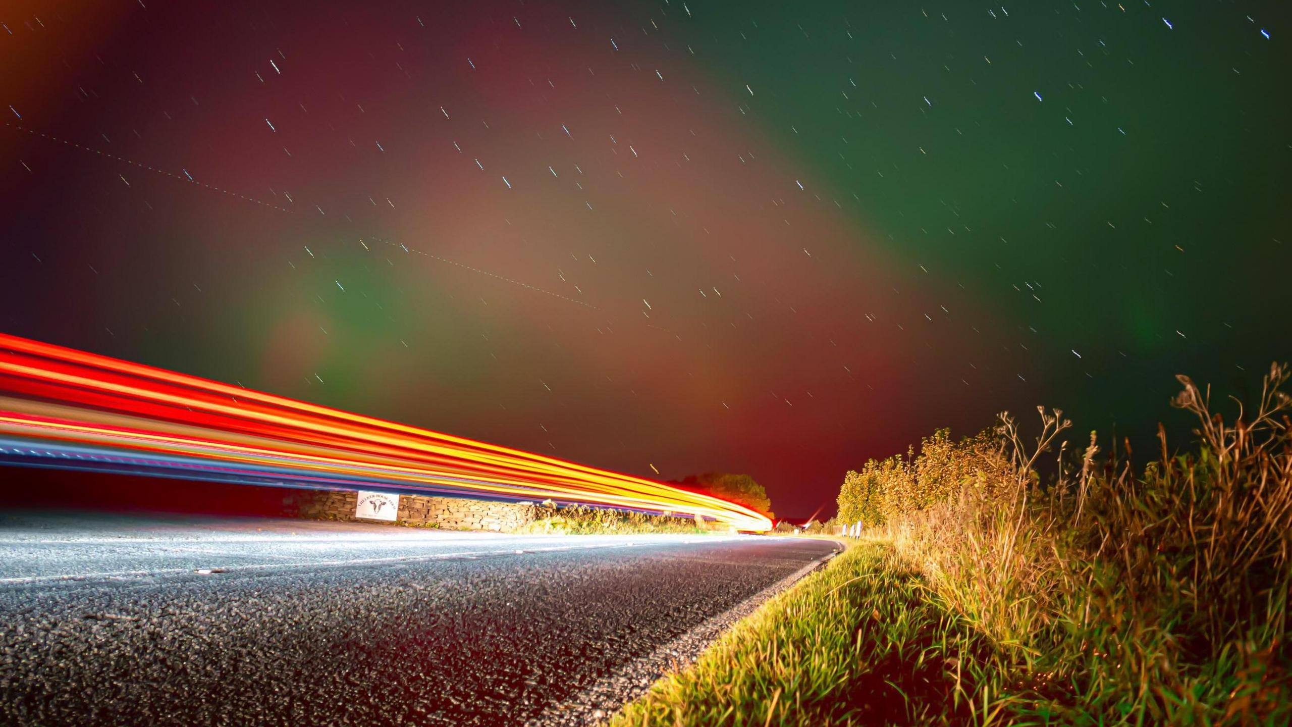 Long exposure shows car lights on a road, with the Northern Lights in the background