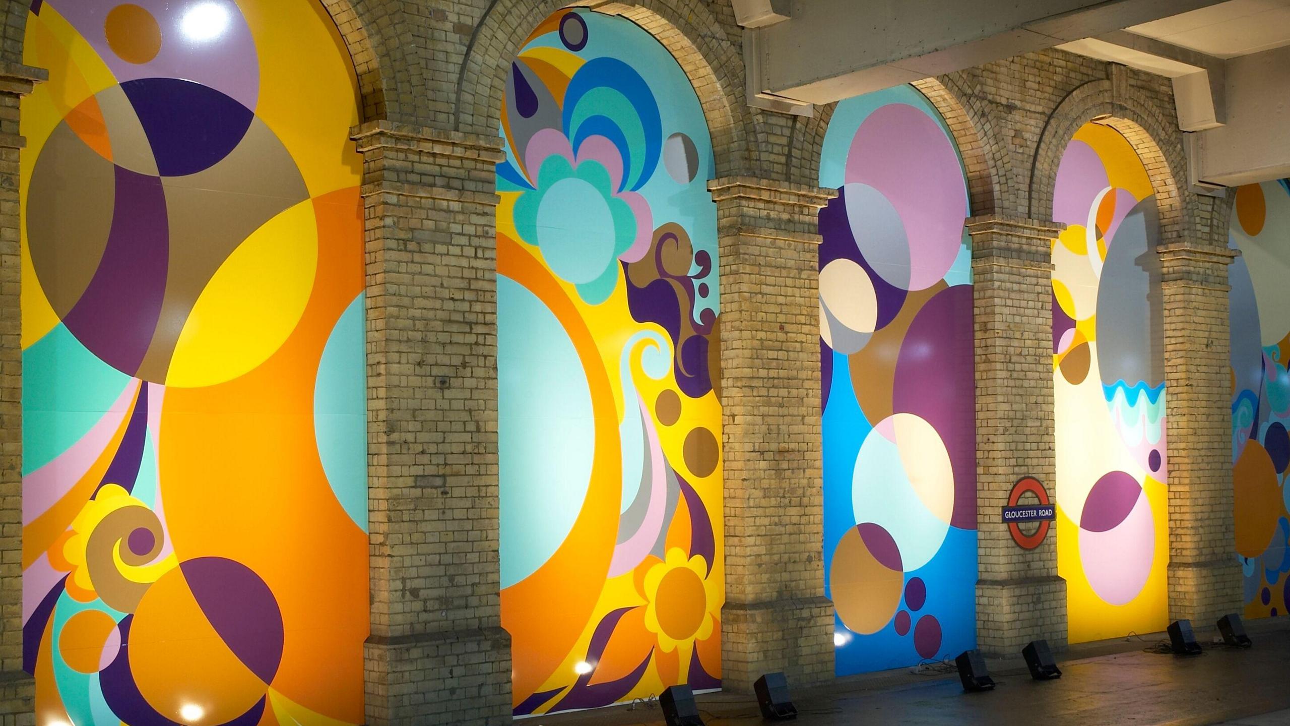 A multi-coloured series of panels run the length of a tube platform, with geometric shapes and motifs. Some represent plants and others represent circles. 