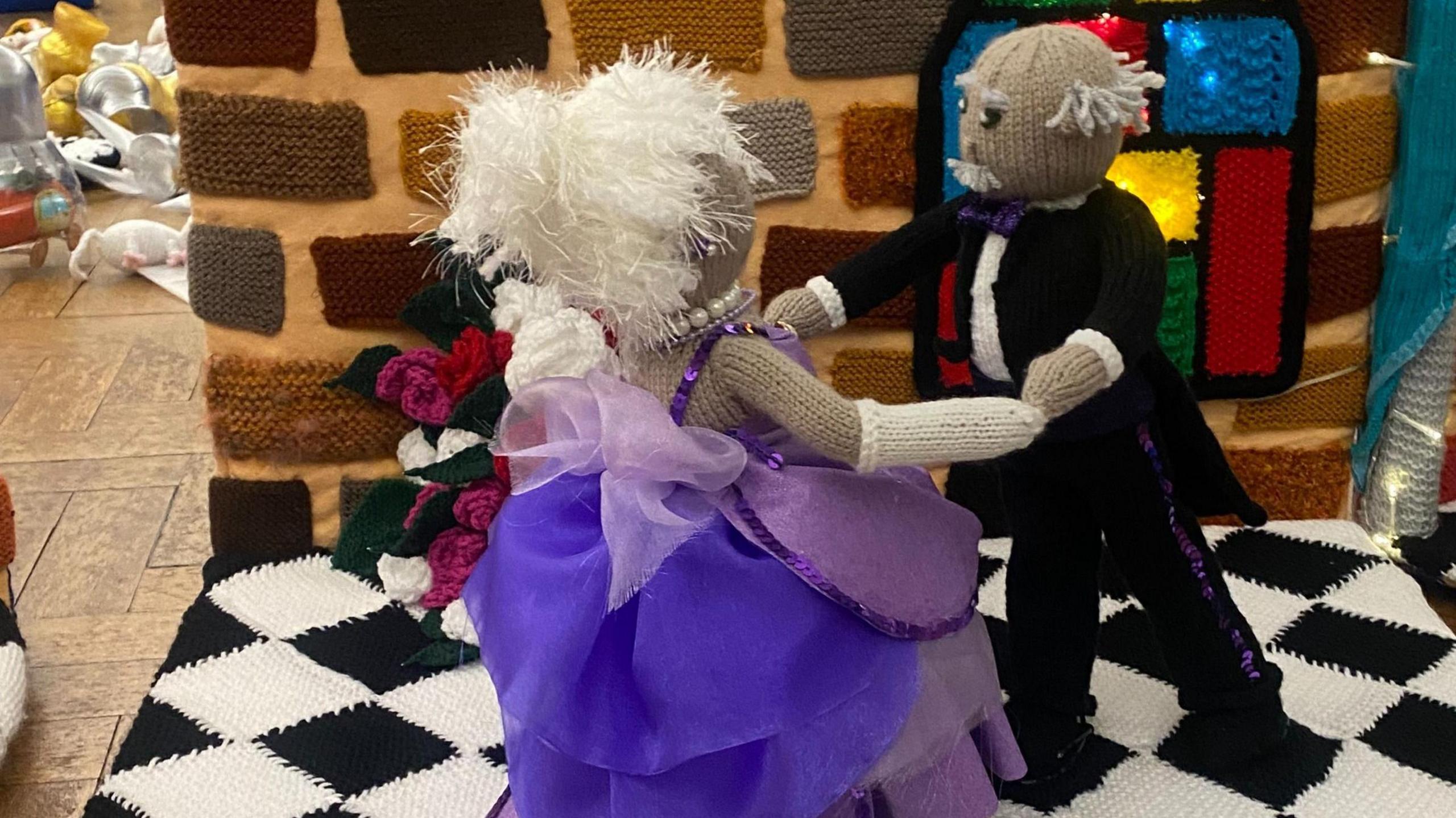 A couple dancing, made out of wool. The woman is wearing a purple dress and the man a black tuxedo. They are on a black and white floor with a knitted brick wall behind them. 