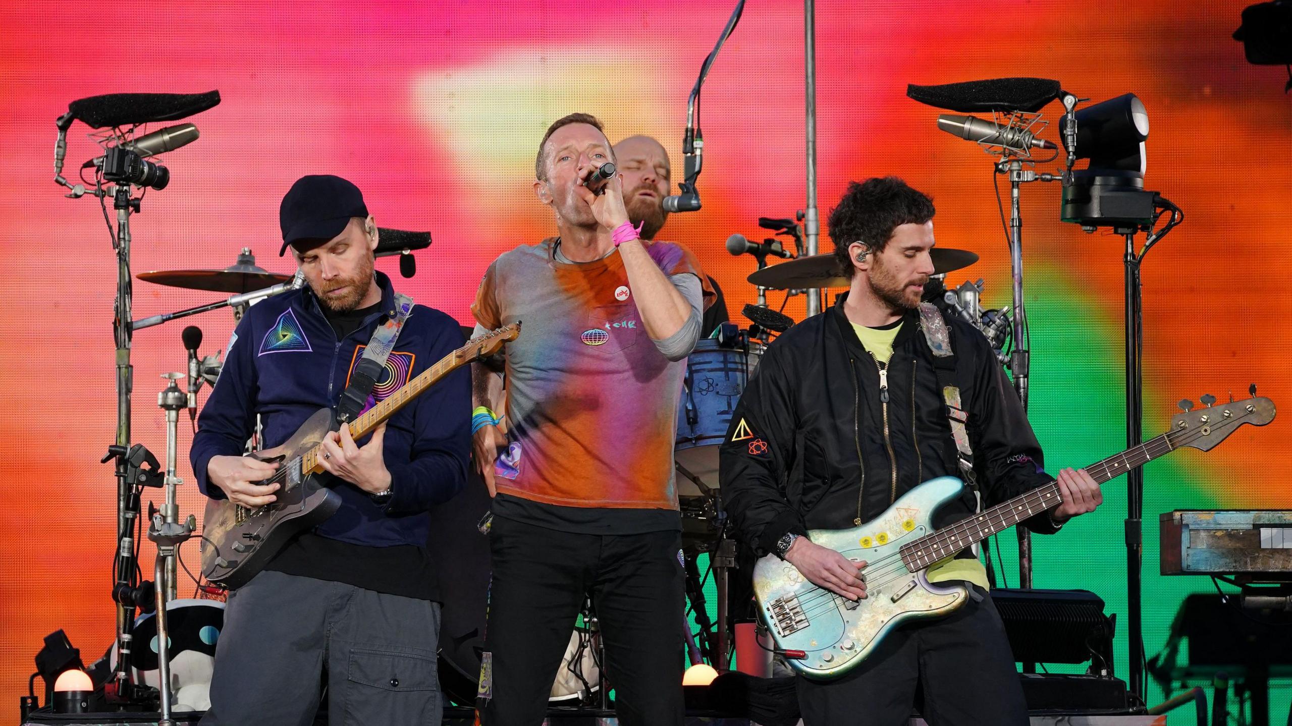 A band performs on stage with three members in the foreground: a guitarist on the left, a singer in the centre, and a bassist on the right. Colourful stage lighting in red, orange, and green fills the background, with drums and equipment visible behind them.